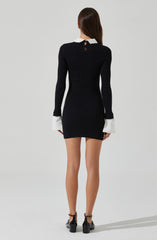 A woman is wearing a fitted black long-sleeve dress with a white collar and white cuffs on the sleeves. The dress has a small keyhole opening at the back neckline with a button closure. She is also wearing black high-heeled shoes with ankle straps.