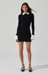 A young woman is wearing a fitted black ribbed dress with long sleeves. The dress features contrasting white shirt cuffs and a white collar. She is also wearing black ankle-strap heels with pointed toes.