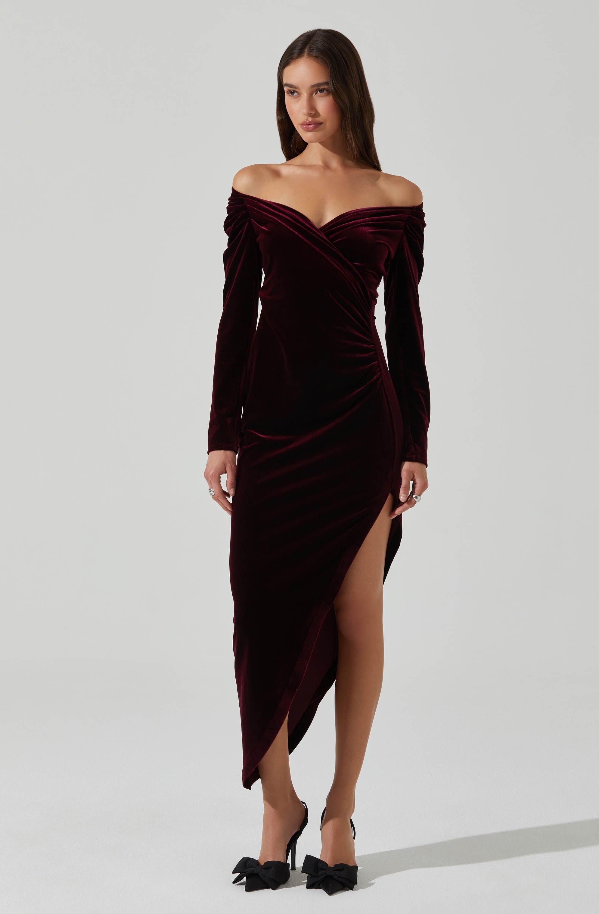 A woman is wearing an off-the-shoulder, long-sleeved dress made of deep burgundy velvet fabric. The dress features a sweetheart neckline with pleated detailing across the bust, a fitted waist, and a high slit on the left side that reveals her leg. She is also wearing black high heels with large bow accents on the toes.