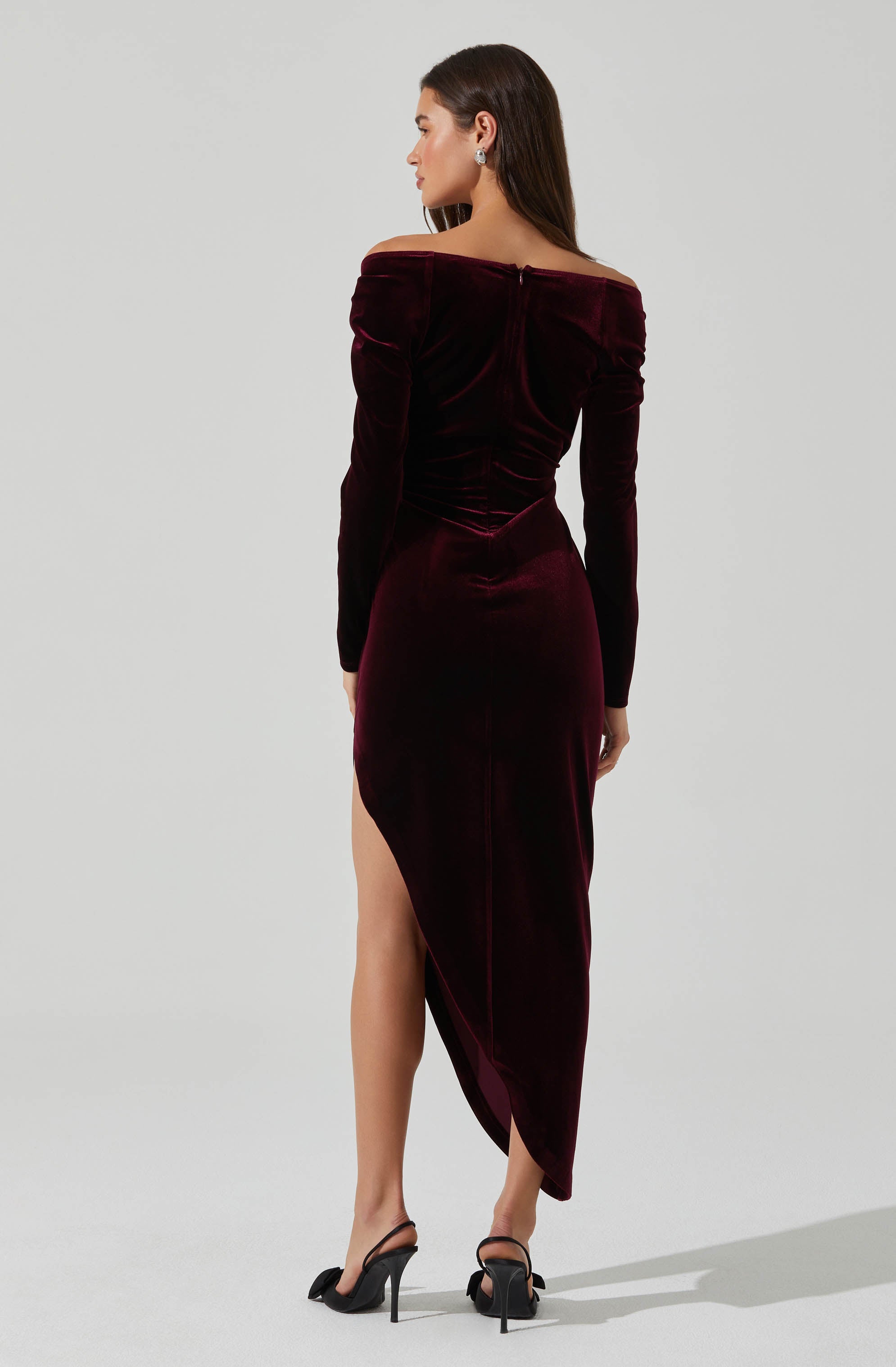 A woman is wearing a long, off-the-shoulder velvet dress in a deep burgundy color. The dress has long sleeves and a fitted silhouette, with a slit on one side revealing her leg. She is also wearing black high-heeled sandals.