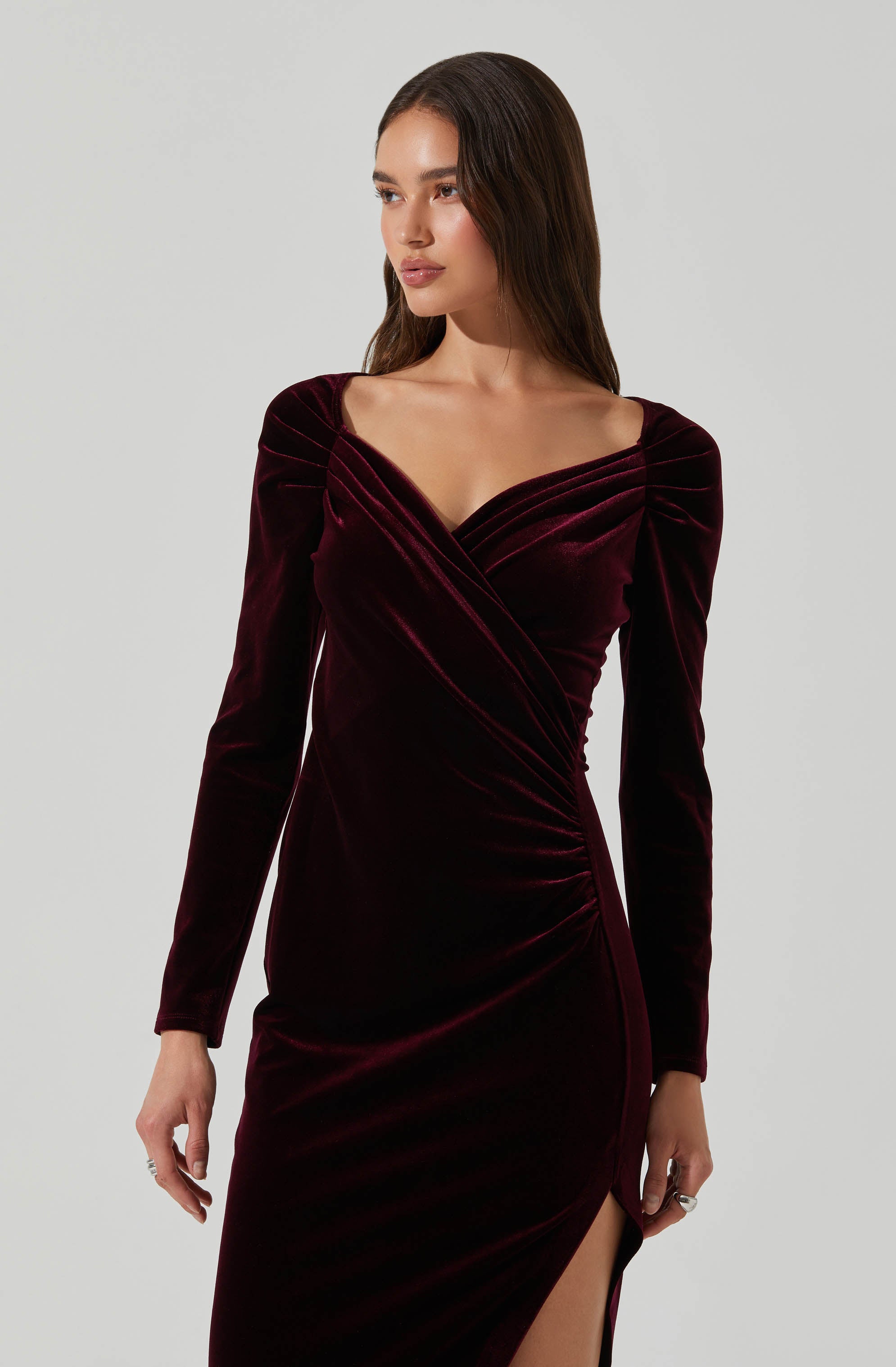 A woman is wearing a long, fitted, dark red velvet dress with long sleeves. The dress features a wrap-style bodice with a deep neckline and gathered detailing at the shoulders and waist, along with a high slit on one side.