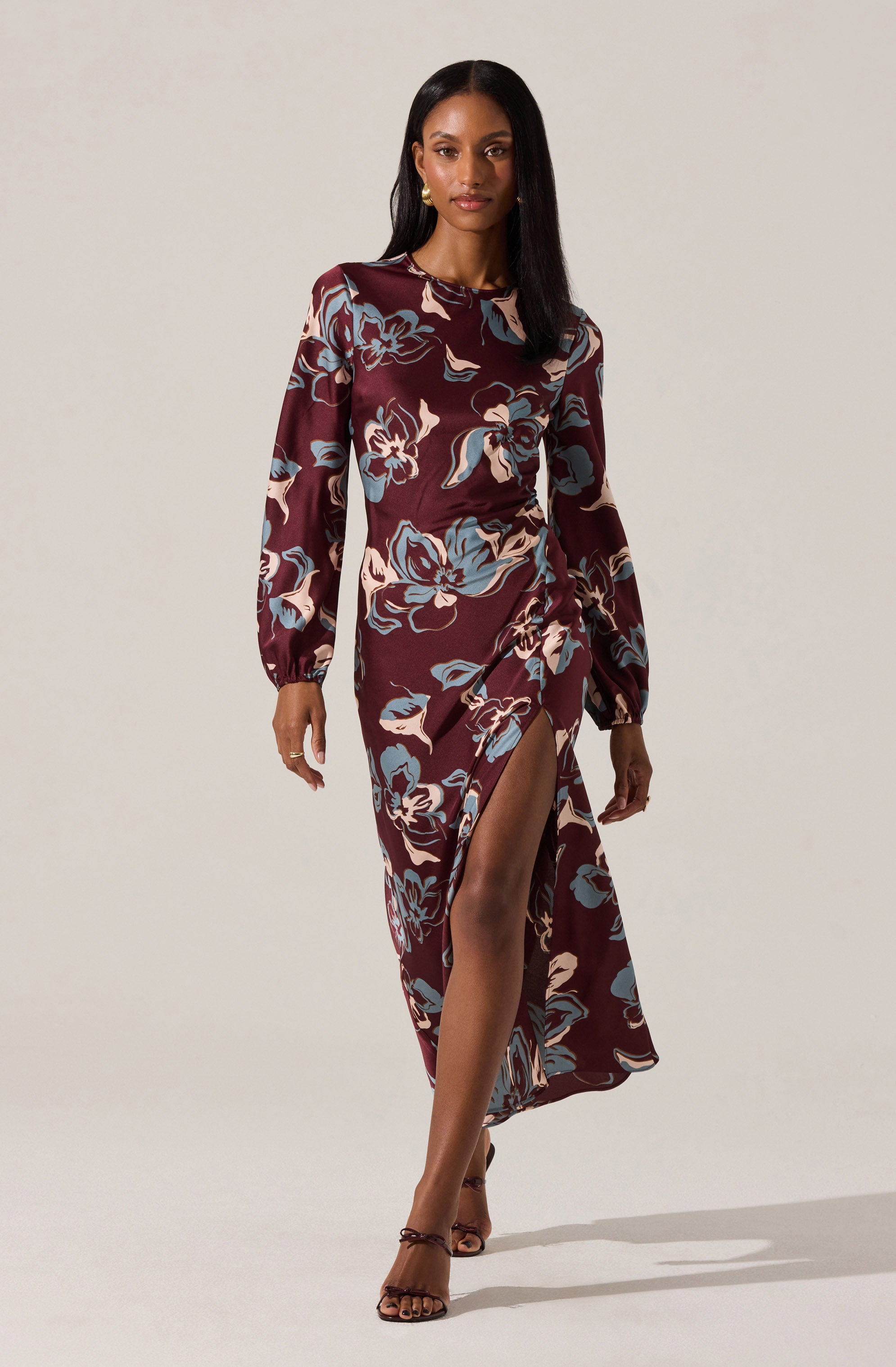 A woman is wearing a long-sleeved, midi-length dress with a high slit. The dress has a dark maroon background with large, abstract floral patterns in shades of blue, cream, and light pink. She is also wearing high-heeled sandals.