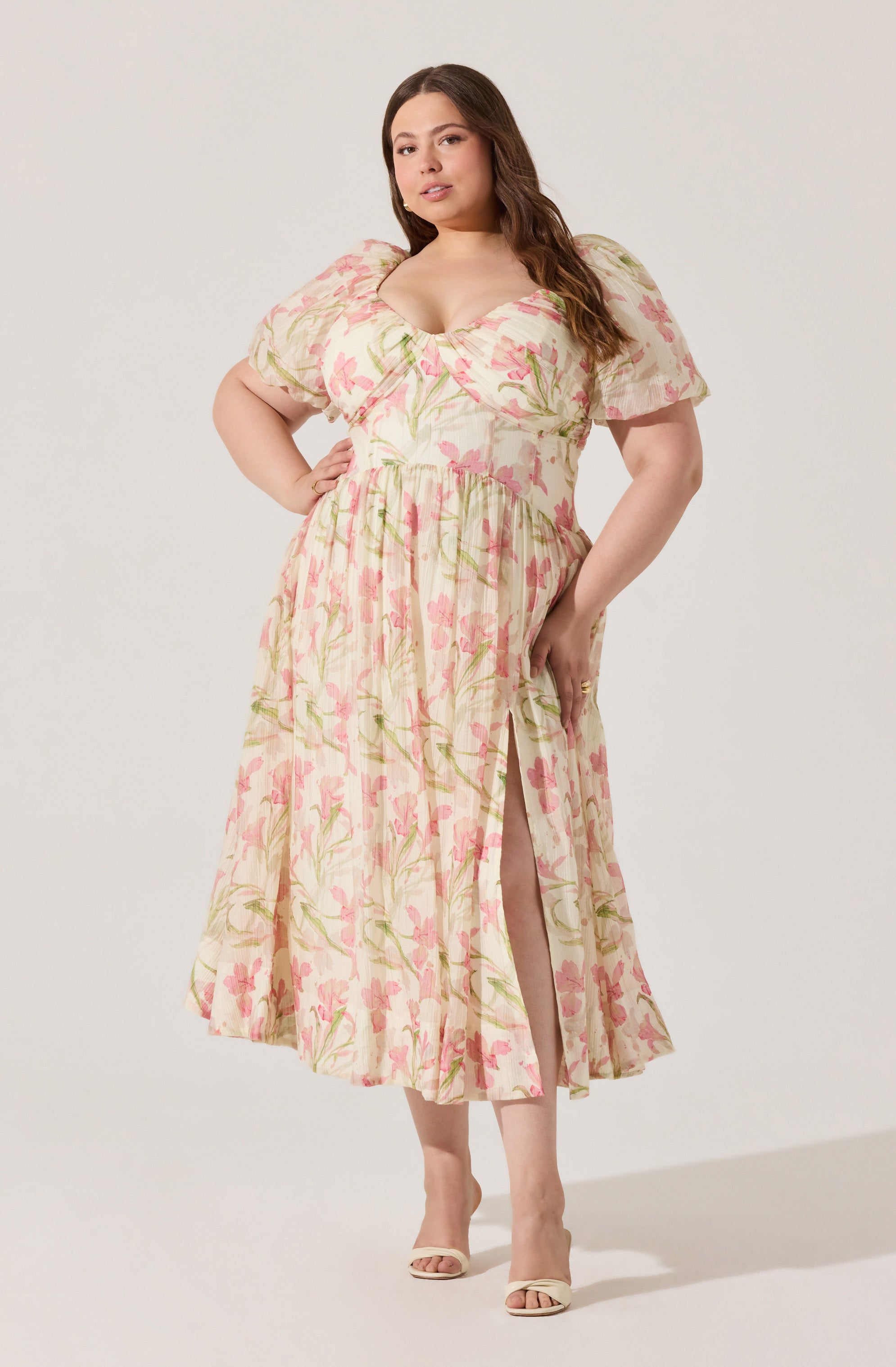 A woman is wearing a long, flowy dress with a light, cream-colored background and a pink and green floral pattern. The dress has short, puffed sleeves, a fitted bodice with a sweetheart neckline, and a high slit in the skirt that reveals her leg. She is also wearing open-toe heels.