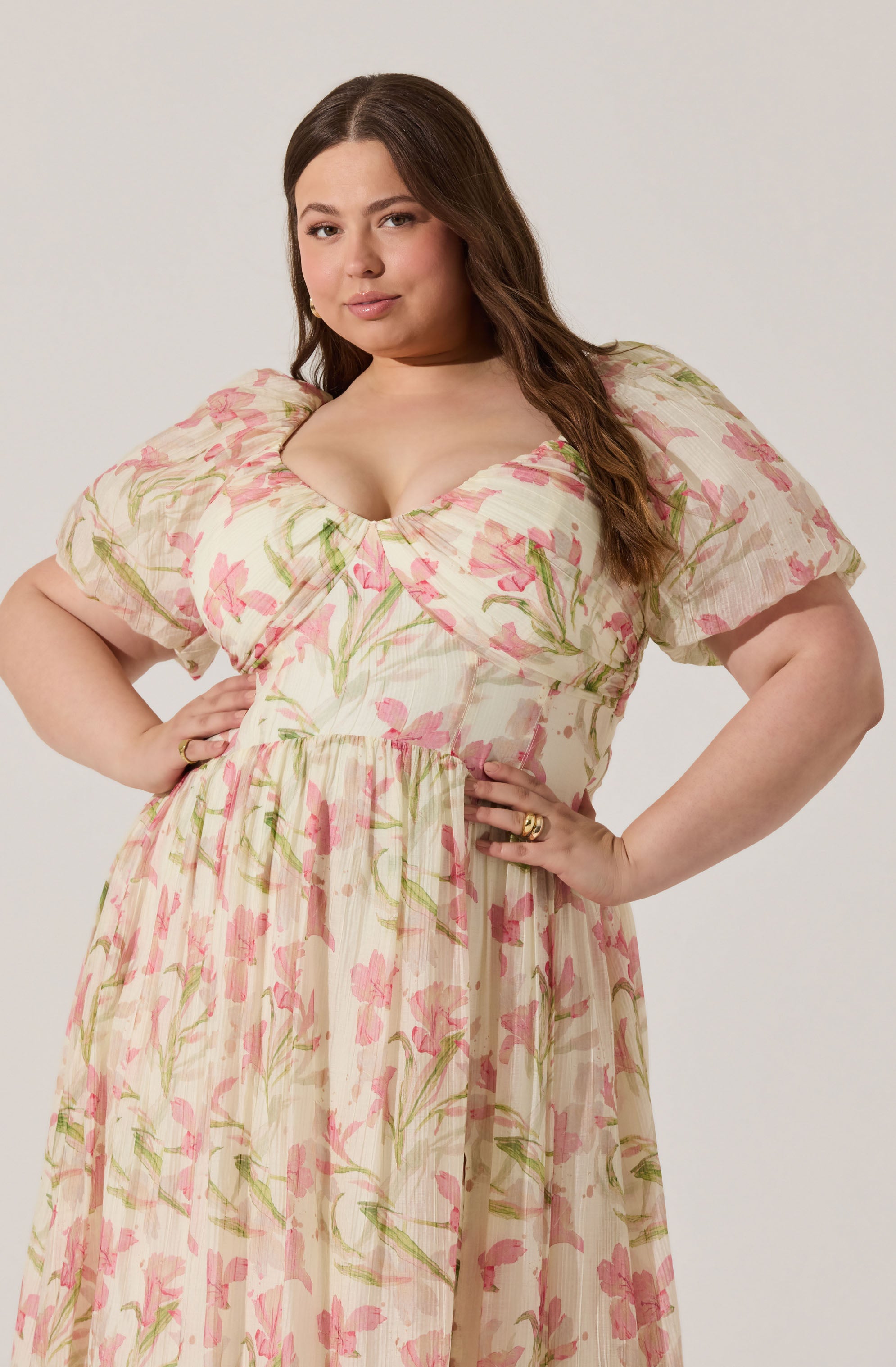 A woman is wearing a flowy, cream-colored dress with pink and green floral patterns. The dress has short, puffed sleeves and a fitted bodice that accentuates her waist, with a gathered skirt that falls to her ankles. The fabric appears lightweight and slightly textured.