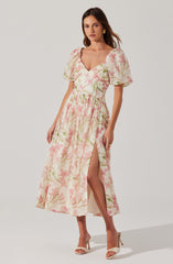 A woman is wearing a long, flowy dress with a pastel pink and green floral print. The dress has a deep V neckline, puffed short sleeves, and a high slit on one side that reveals her leg. She is also wearing beige high-heeled sandals and gold earrings.