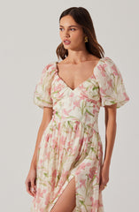 A woman is wearing a light-colored dress with a pink and green floral print. The dress features puffed short sleeves, a sweetheart neckline, and a gathered waist. The fabric appears to be lightweight and textured, with a slit on the leg revealing her leg. She has long, wavy brown hair and is accessorized with gold earrings.