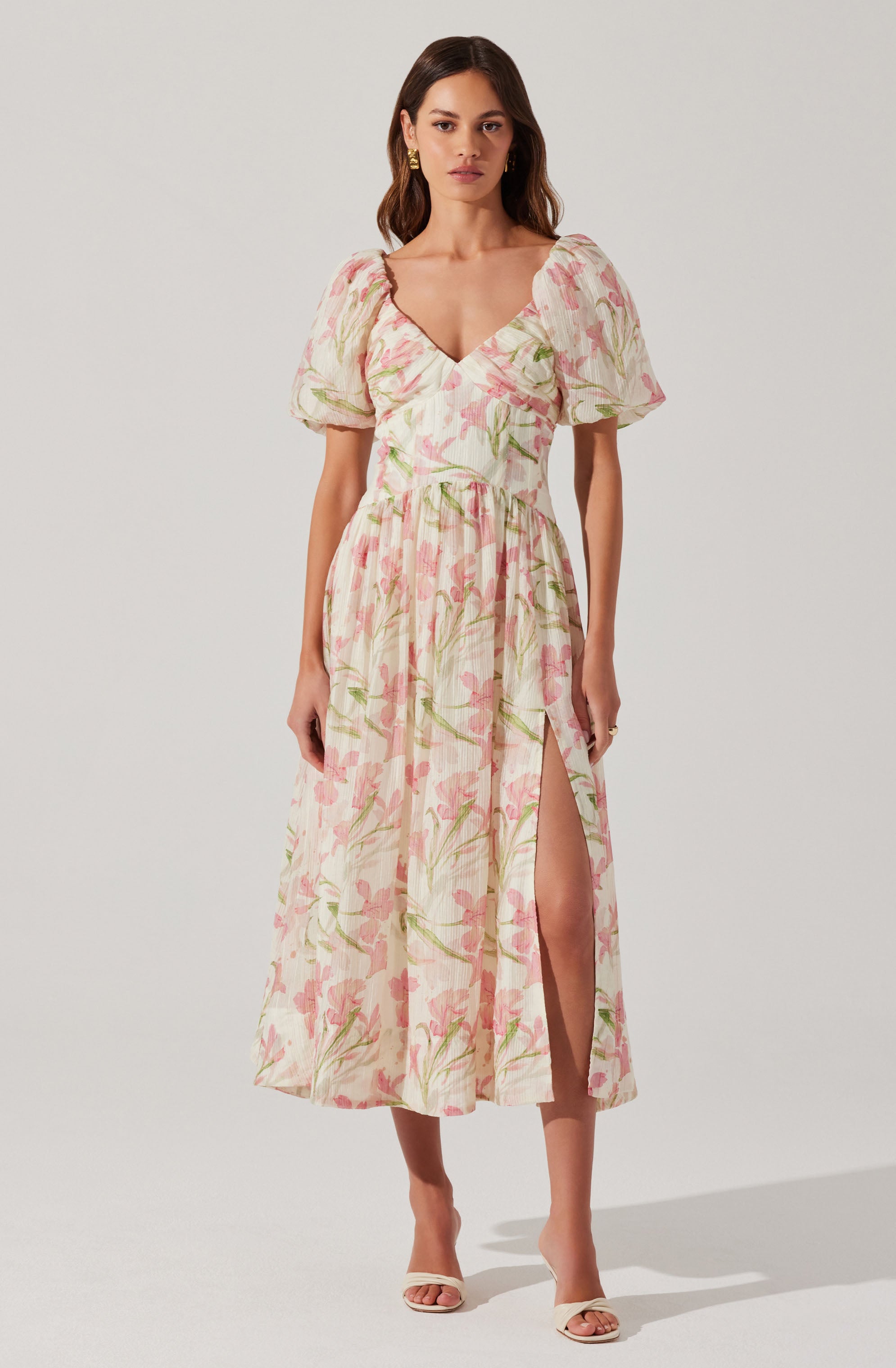 A woman is wearing a long, flowy dress with a floral pattern featuring pink and green colors on a light background. The dress has short, puffed sleeves and a deep V-neckline. It also has a high slit on one side, revealing her leg.