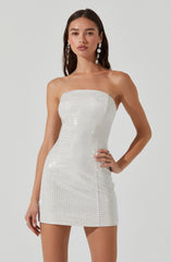 A woman is wearing a strapless, fitted mini dress made of a shiny, white fabric that appears to be embellished with small, sparkling beads or sequins. The dress is sleek and form-fitting, ending high on the thighs. She has long dark hair and is accessorized with large, dangling earrings.