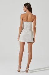 A woman is wearing a strapless, fitted, cream-colored mini dress with a subtle shimmer. She has on matching high-heeled sandals with ankle straps. Her accessories include large statement earrings.