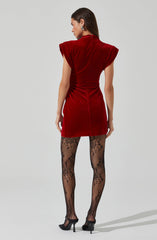 A woman is wearing a red velvet dress with structured, padded shoulders. She has on patterned black lace tights and black high-heeled shoes. She is also accessorized with large, statement earrings.