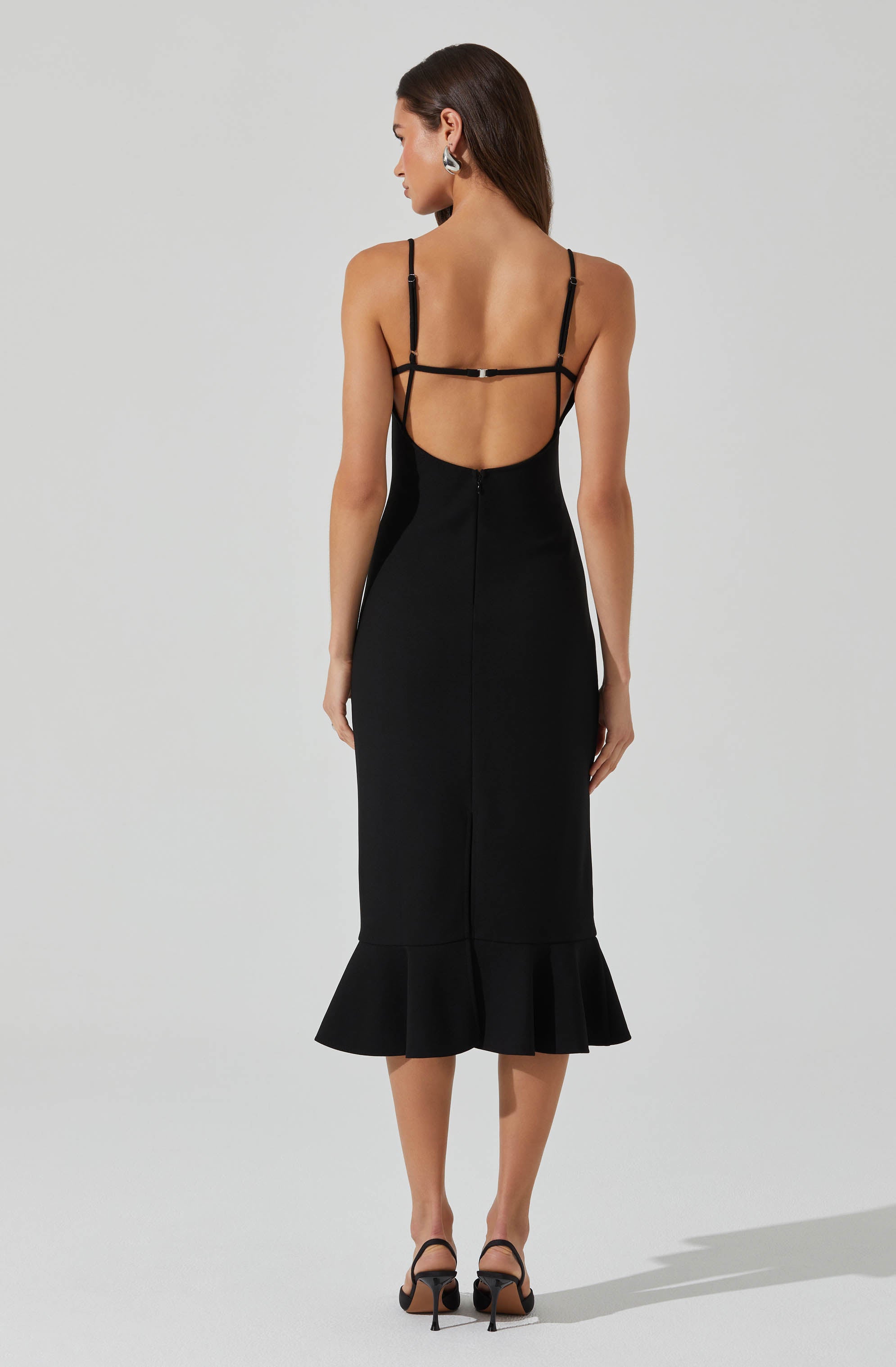 A woman is wearing a black, sleeveless, midi-length dress with thin spaghetti straps. The dress features an open back with multiple black straps across the upper back and shoulders, creating a strappy design. The lower part of the dress has a ruffled hem, adding a flouncy detail to the bottom.