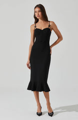 A woman is wearing a form-fitting black dress with thin adjustable straps that have small bows at the shoulders. The dress has a fitted bodice with seam detailing and a flared ruffle hem at the bottom. She is also wearing black high-heeled shoes with a decorative element on the toe.