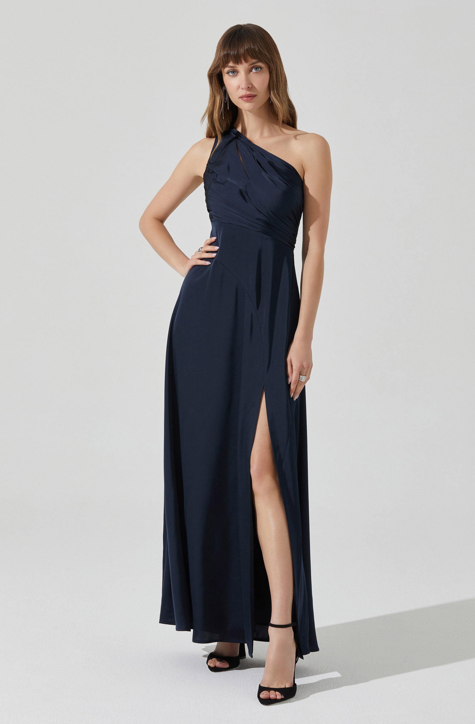 A woman is wearing a floor-length navy blue evening gown with an asymmetrical, one-shoulder neckline. The dress features gathered fabric at the bodice and a high slit on one side, revealing her leg. She is wearing black high-heeled sandals and minimal jewelry.