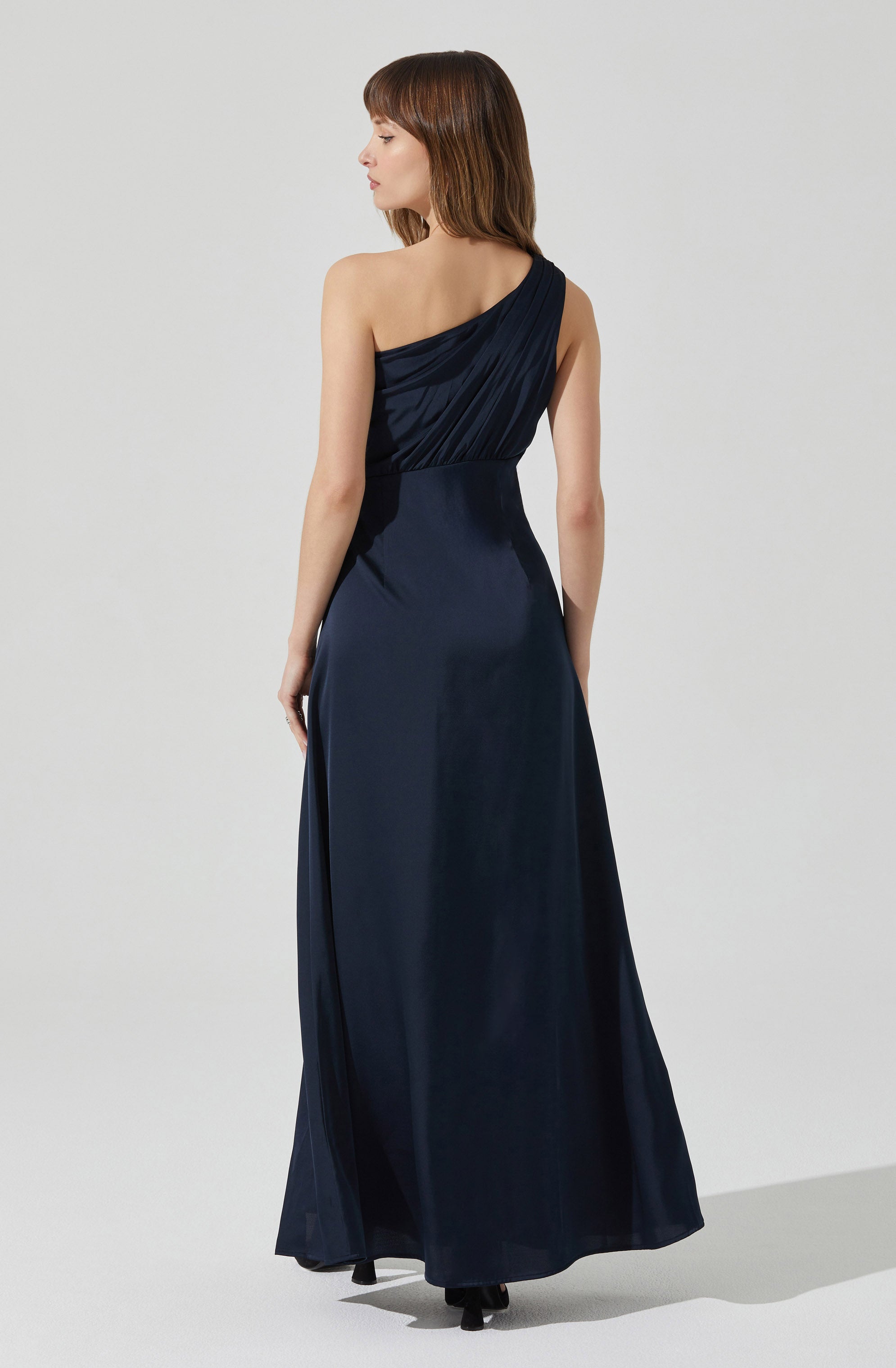 A woman is wearing a floor-length, navy blue evening gown. The dress features a one-shoulder design with a draped, ruched bodice. The fabric appears to be satin or silk, giving it a smooth and shiny finish.