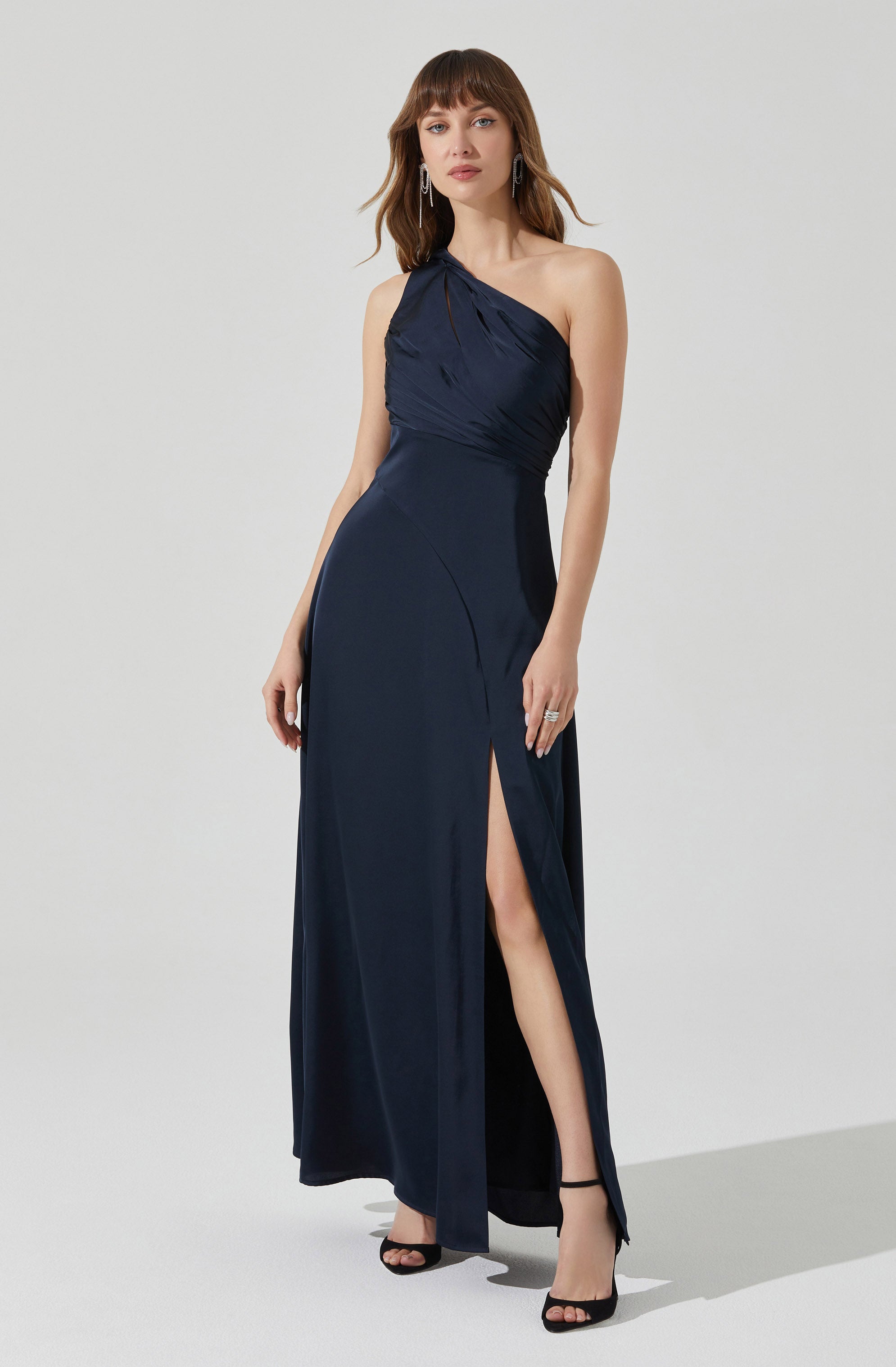 A woman is wearing an elegant, dark blue, one-shoulder evening gown with a fitted bodice featuring gathered fabric details. The dress has a thigh-high slit on one side, revealing her leg and black high-heeled shoes. She accessorizes with large dangling earrings and rings.