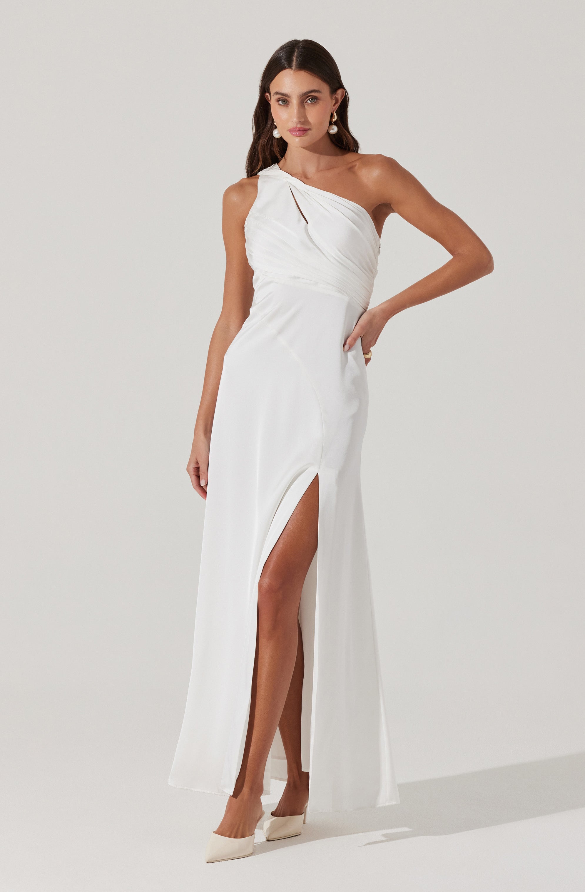 A woman is wearing an elegant white evening gown with a one-shoulder design. The dress features draped fabric across the bust and waist, with a high slit on one side revealing her leg. She is accessorized with large pearl earrings and cream-colored high heels.