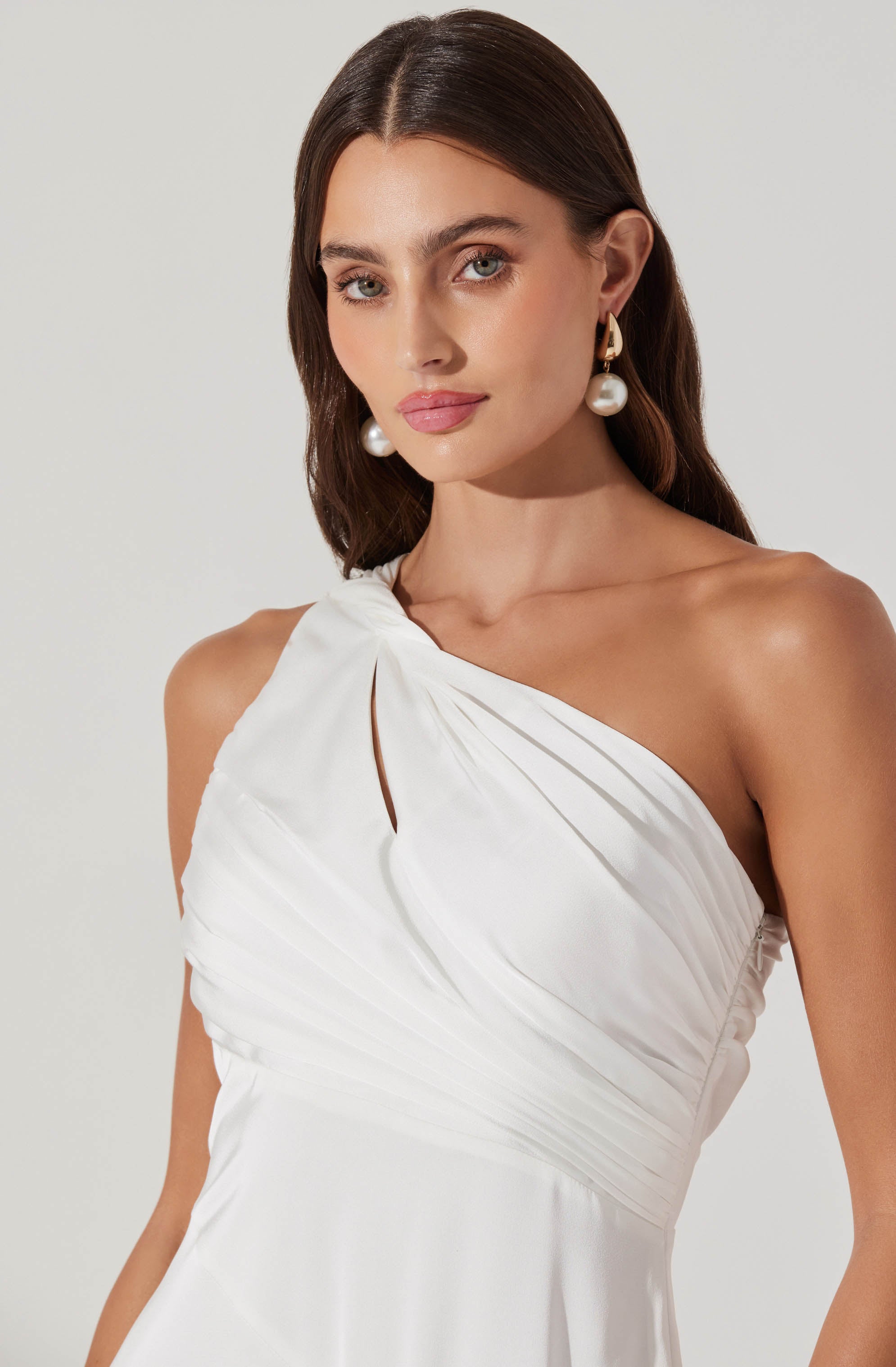 A woman is wearing a white, one-shoulder dress with pleated fabric. The dress has a gathered design at the shoulder, creating a slightly draped effect across the bust. She accessorizes with large pearl earrings and has long, dark brown hair styled in soft waves.