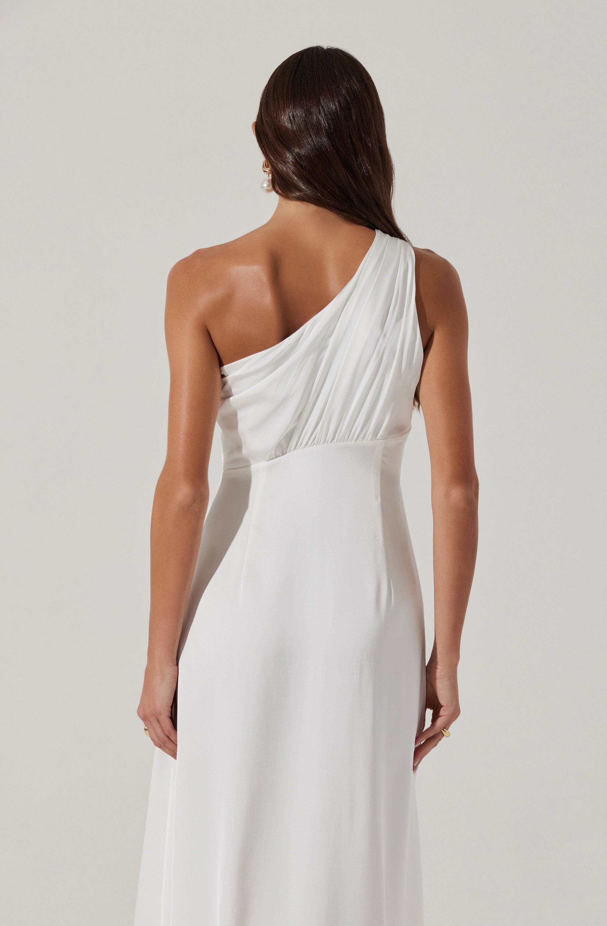 The dress is a sleeveless, floor-length gown in a smooth, white fabric. It features an asymmetrical, one-shoulder design with pleated detailing on the shoulder strap that drapes diagonally across the chest. The back of the dress is fitted and sleek, emphasizing a streamlined silhouette.