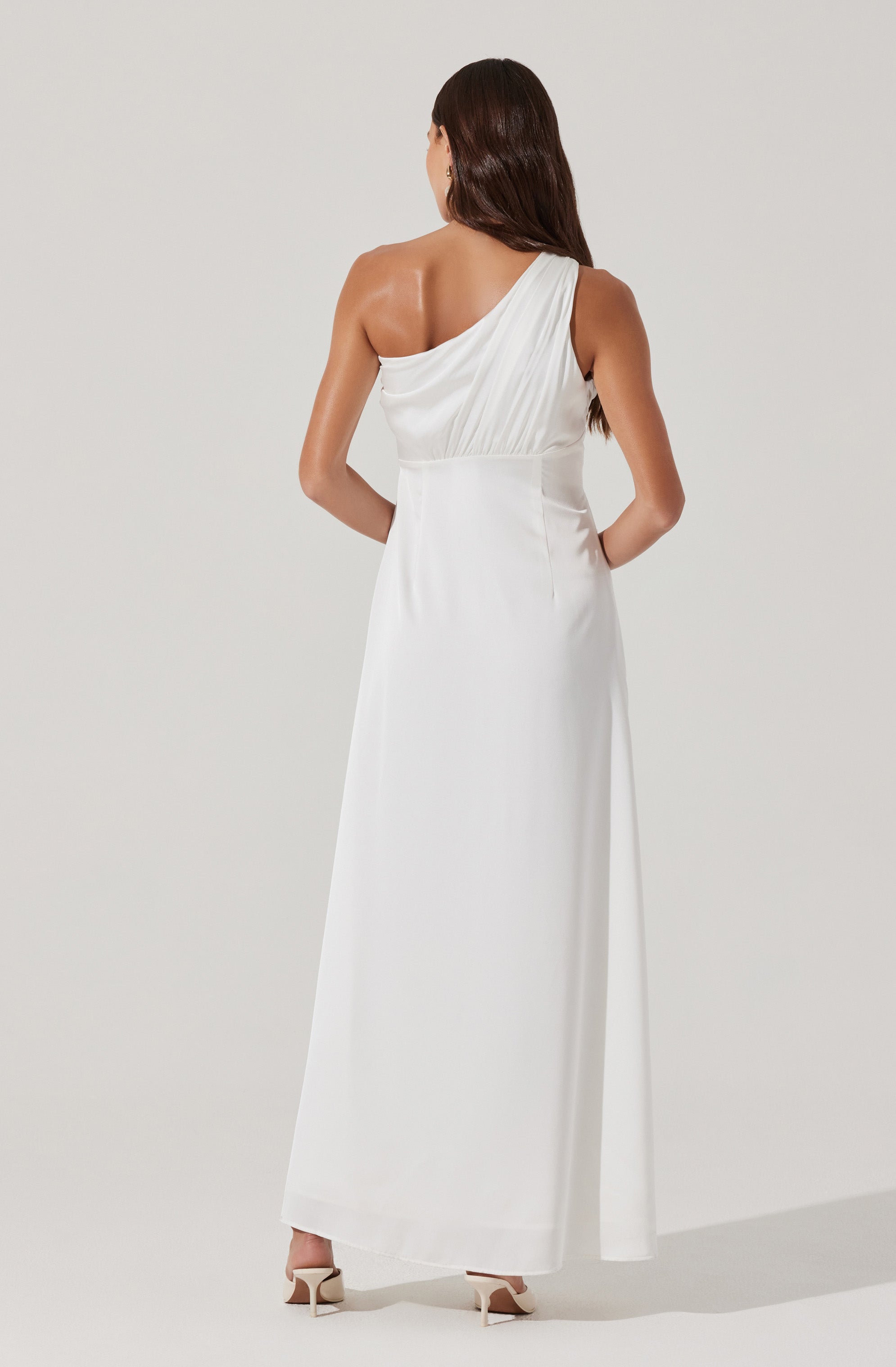 A woman is wearing a long, elegant white dress with an asymmetrical neckline and draped detailing on the bodice. The dress extends to the floor, creating a smooth, flowing silhouette. She is also wearing nude high-heeled shoes.