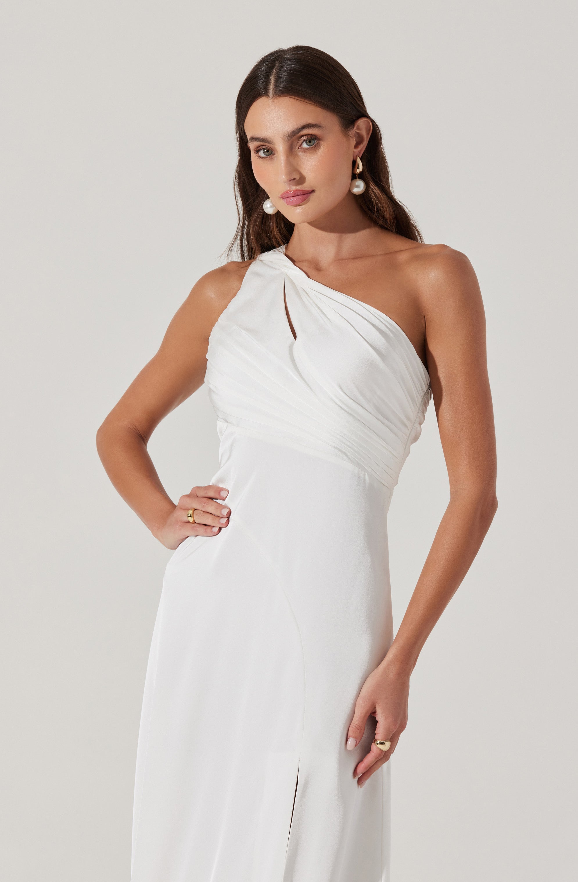A woman wearing an elegant, one-shoulder white gown with pleated fabric across the bodice. The dress has a small keyhole cutout near the shoulder and a high slit at the front. She accessorizes with large pearl earrings and rings on her fingers.