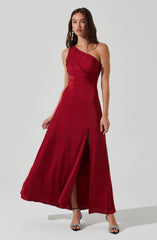 A woman is wearing a floor-length, one-shoulder red gown with ruched detailing on the bodice. The dress features a high slit in the front, revealing her legs. She pairs the gown with open-toe high heels and statement earrings.