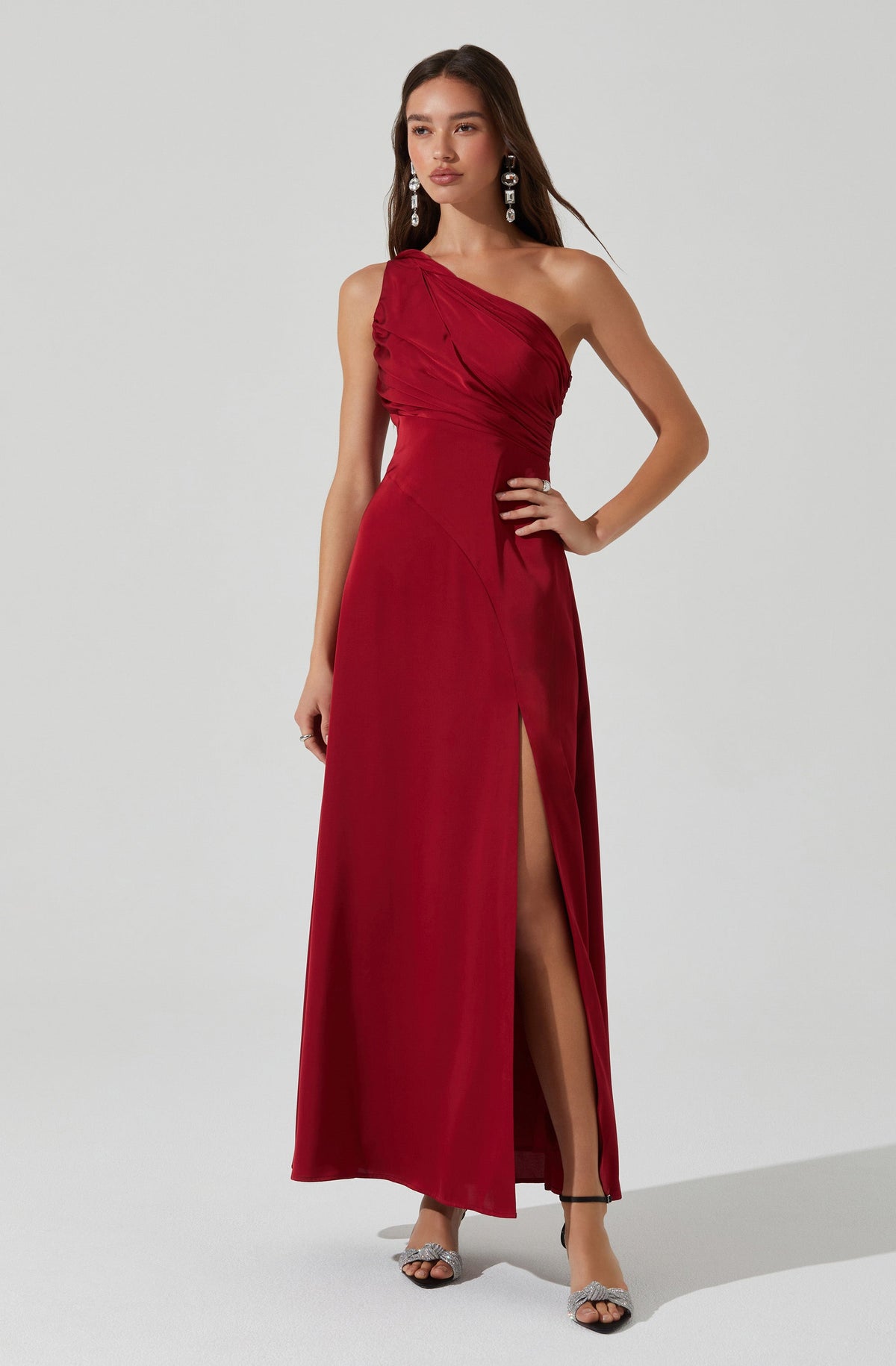 A woman is wearing a long, elegant, strapless red dress. The dress features a gathered, ruched bodice on one side and a high slit on the front left side, revealing her leg. She accessorizes with large, dangling earrings and open-toed heels with ankle straps.