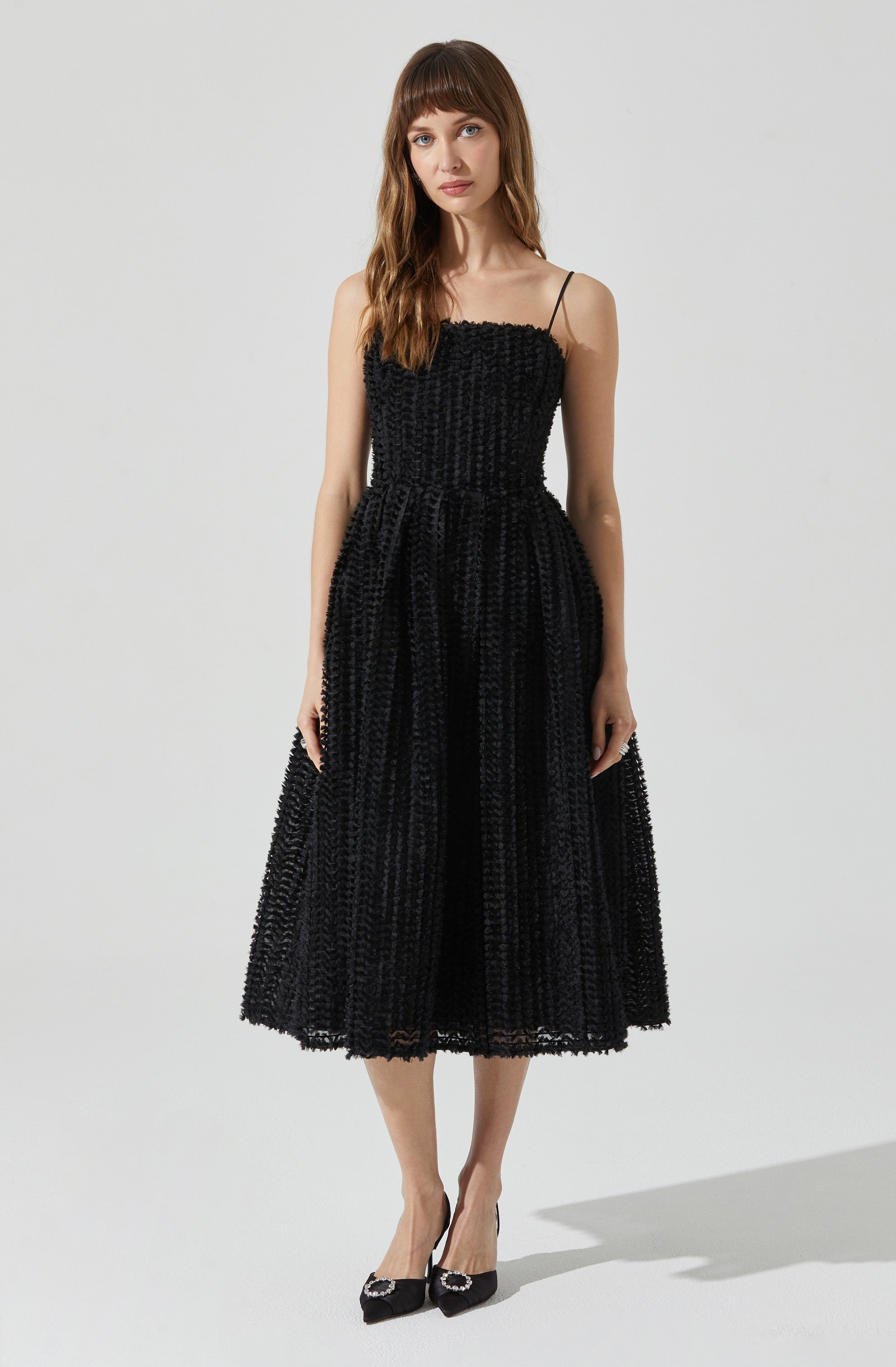 A woman is wearing a black dress with a textured, button-like pattern. The dress has thin spaghetti straps, a fitted bodice, and a full, midi-length skirt. She is also wearing black high heels with embellishments on the toes.