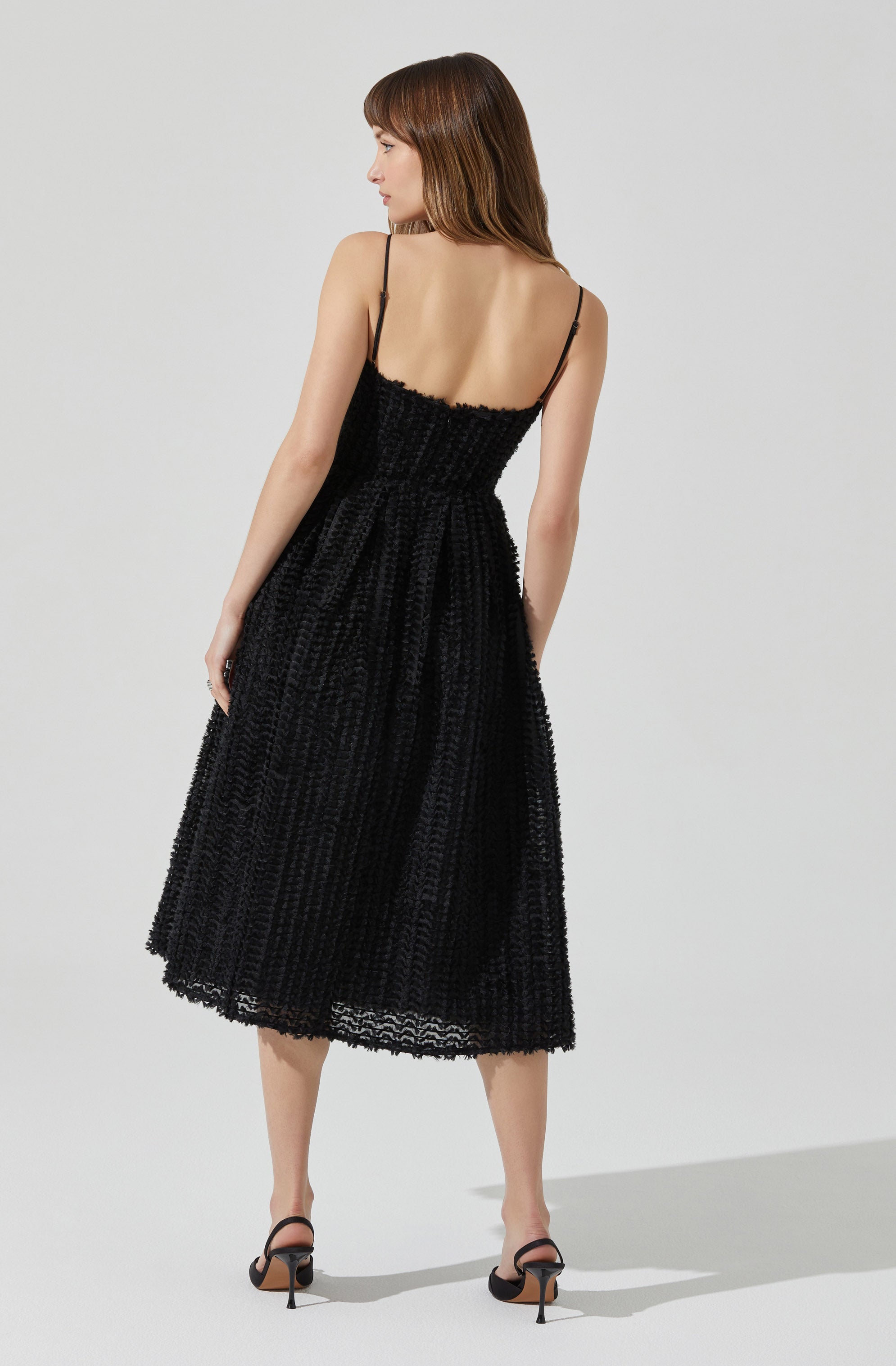 A woman is wearing a black, textured, sleeveless midi dress with thin spaghetti straps. The dress has a fitted bodice and a full skirt, featuring a woven or crochet-like pattern. She is also wearing black high-heeled sandals.