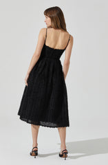 A woman is wearing a black, textured, sleeveless midi dress with thin spaghetti straps. The dress has a fitted bodice and a full skirt, featuring a woven or crochet-like pattern. She is also wearing black high-heeled sandals.