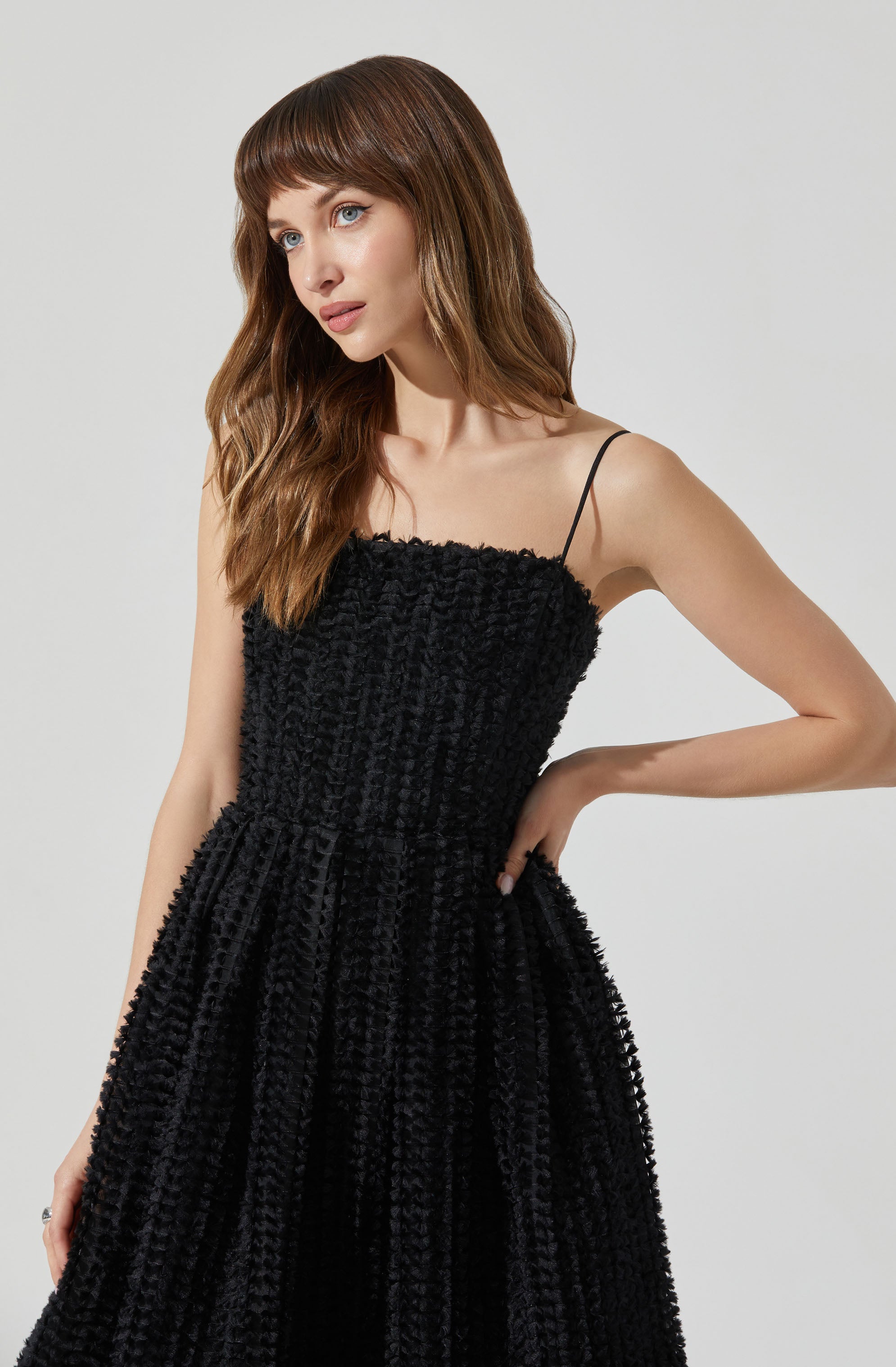 A woman is wearing a black dress with textured, ruched fabric. The dress features thin spaghetti straps, a fitted bodice, and a full skirt that flows downward.