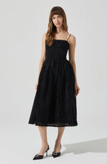 A woman is wearing a black, textured, sleeveless dress with thin spaghetti straps. The dress has a fitted bodice and a full, midi-length skirt with a waffle-like pattern. She pairs the dress with black pointed-toe high heels that have decorative embellishments on the front.