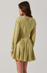 A woman is wearing a light olive-green dress with long sleeves. The dress features a fitted waist with pleats that create a flared, skirt-like bottom. The fabric appears to be smooth and slightly shiny. The back of the dress has a center seam running from the neckline to the waist.
