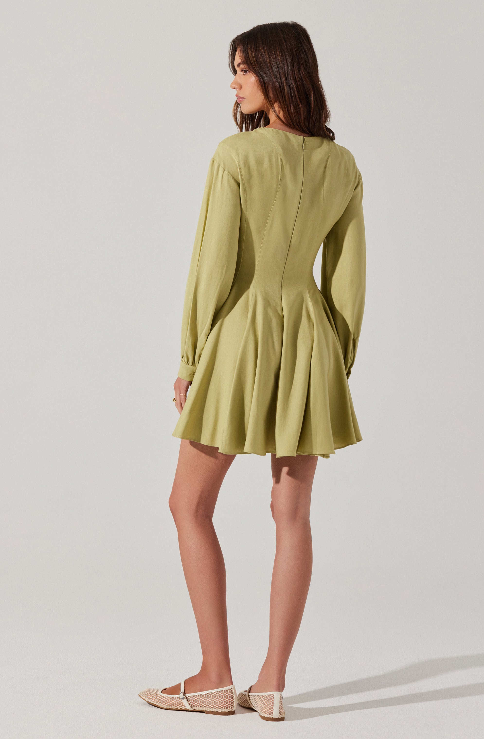A woman is wearing a light green dress with long sleeves, which features a fitted bodice and a flared, pleated skirt that falls above the knees. The dress has a subtle seam detail running down the back. She is also wearing beige woven flats.