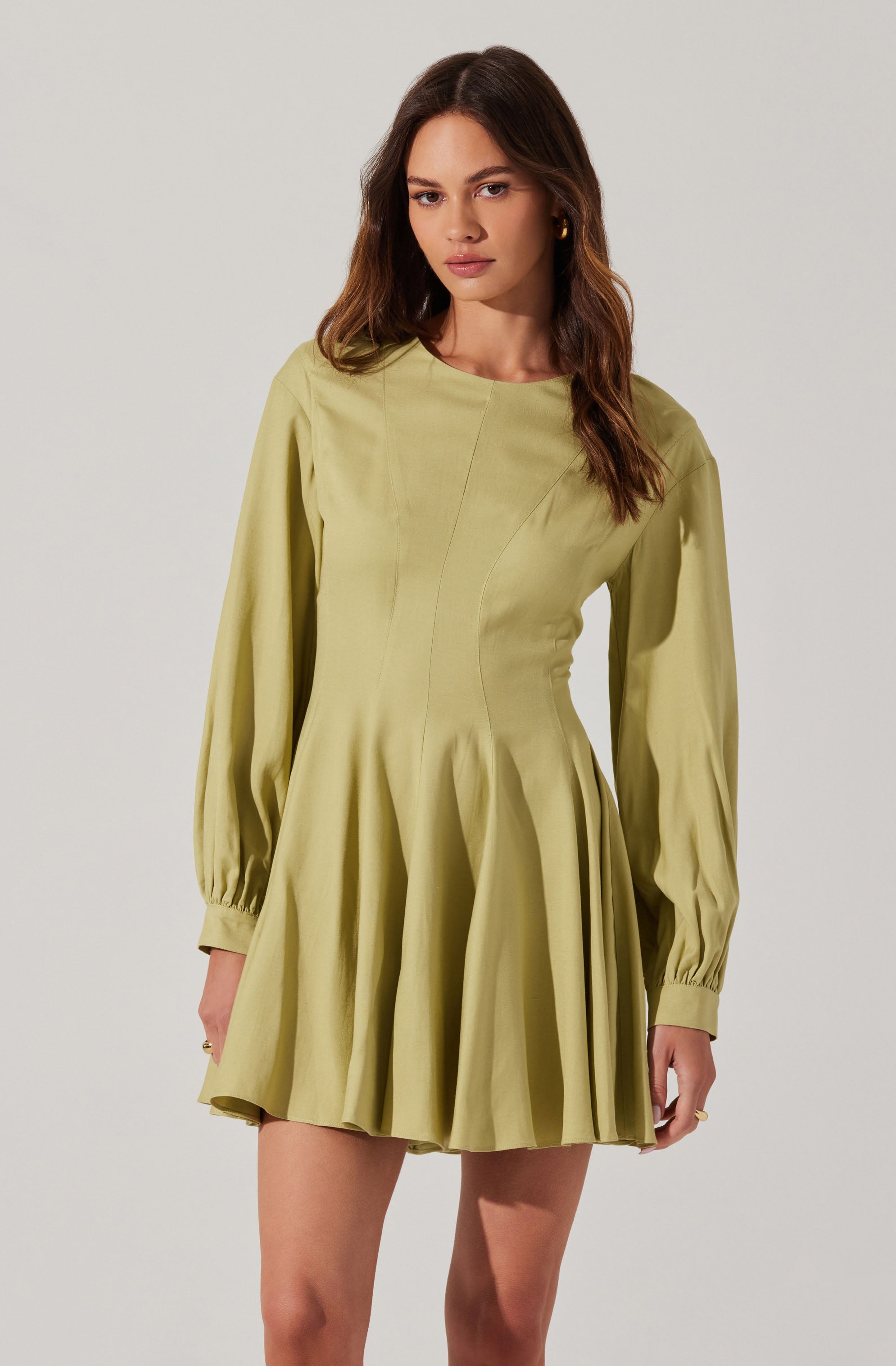 A woman is wearing a light green dress with long, puffed sleeves that are gathered at the wrists with cuffs. The dress has a fitted bodice with vertical seam details and a flared, pleated skirt that falls above the knees. The fabric appears to be smooth and slightly shiny.