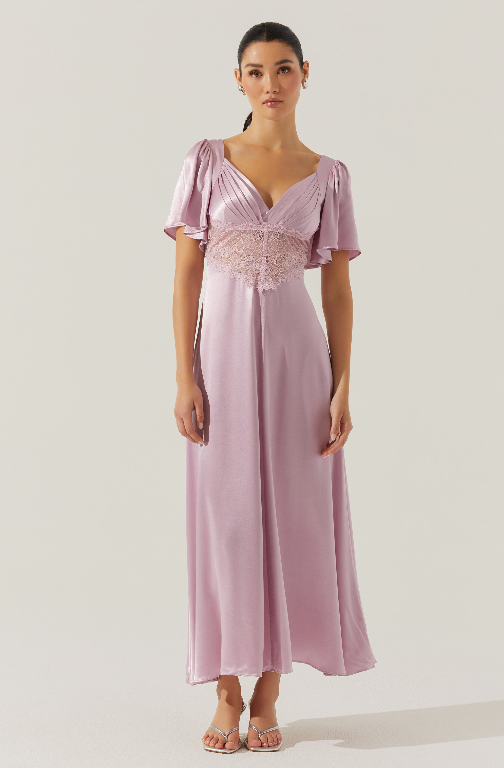 A woman is wearing a long, elegant lavender dress with a fitted bodice and flowing skirt. The dress features short, puffed sleeves with gathered fabric at the shoulders, a v-neckline, and a lace band around the waist. The fabric appears silky and smooth, with a soft sheen. She is also wearing strappy high-heeled sandals.