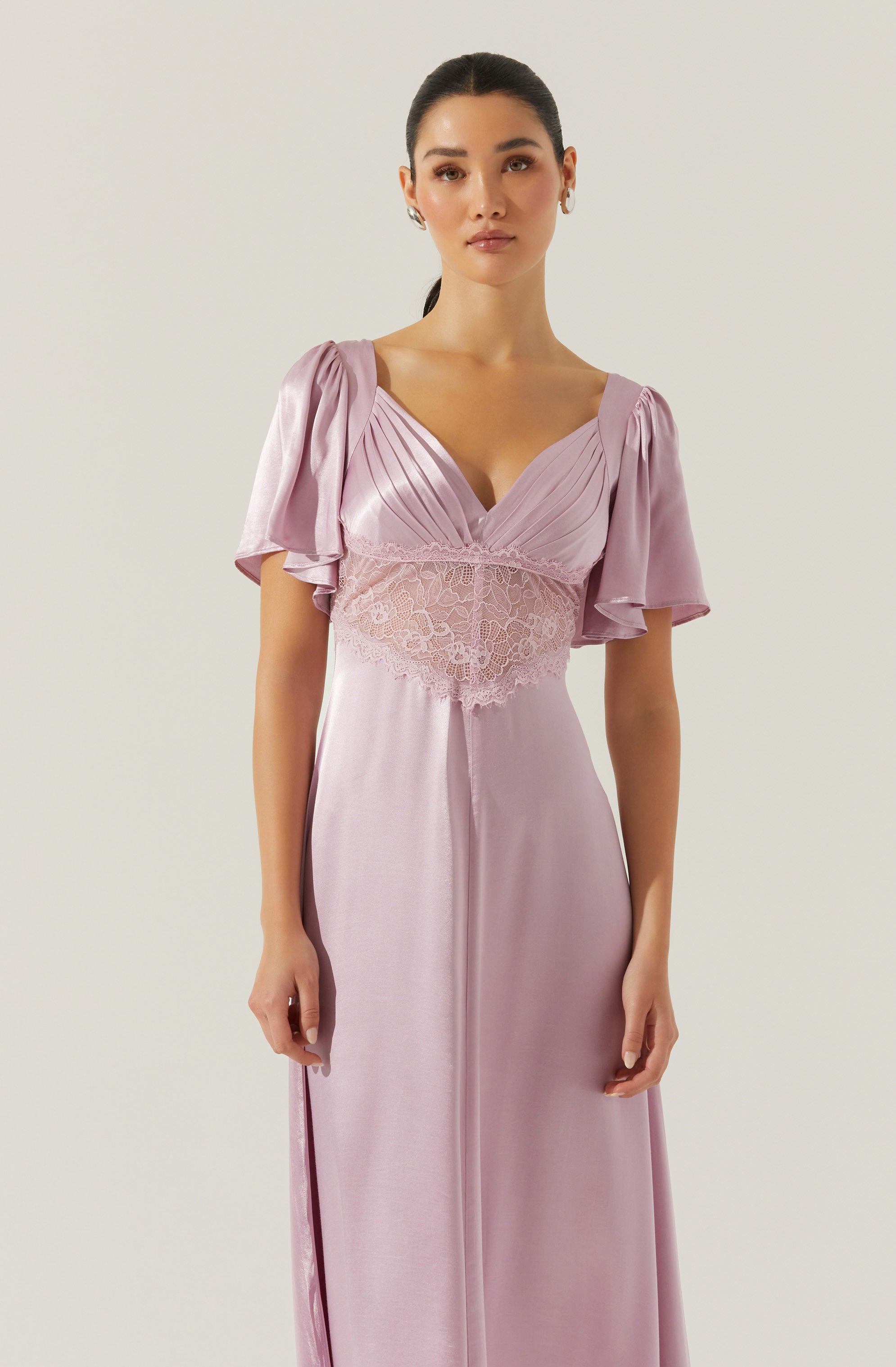 A woman is wearing a floor-length, pastel pink dress with short, puffed sleeves made of a shiny, satin-like fabric. The dress features a V-neckline and a lace band around the waist with floral embroidery, creating a fitted silhouette.