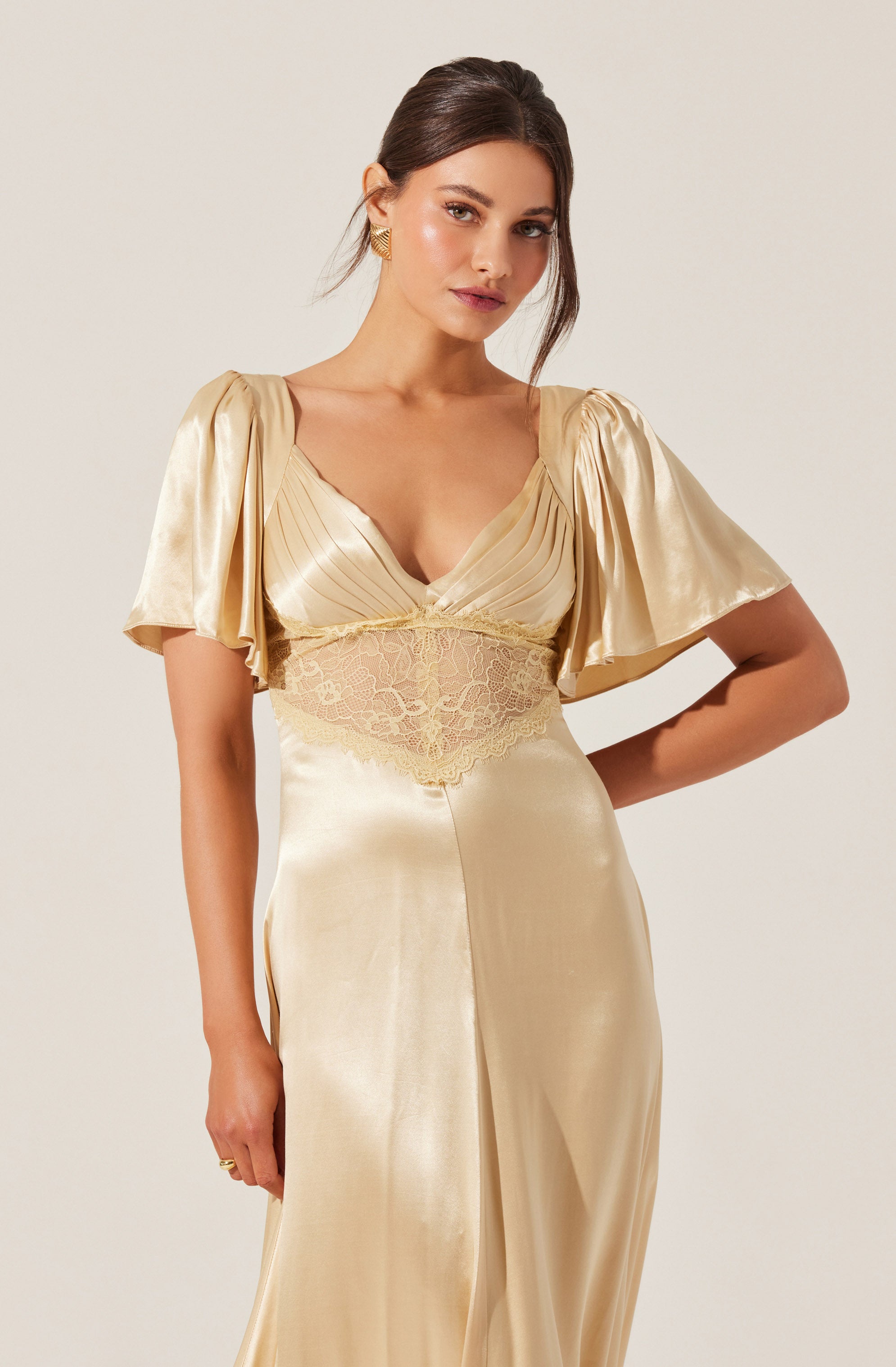 A woman is wearing a satin gown in a champagne color. The dress features short, puffed sleeves and a fitted bodice with pleated fabric and lace detailing at the waist. The skirt is smooth and flowing, with a subtle slit at the front.