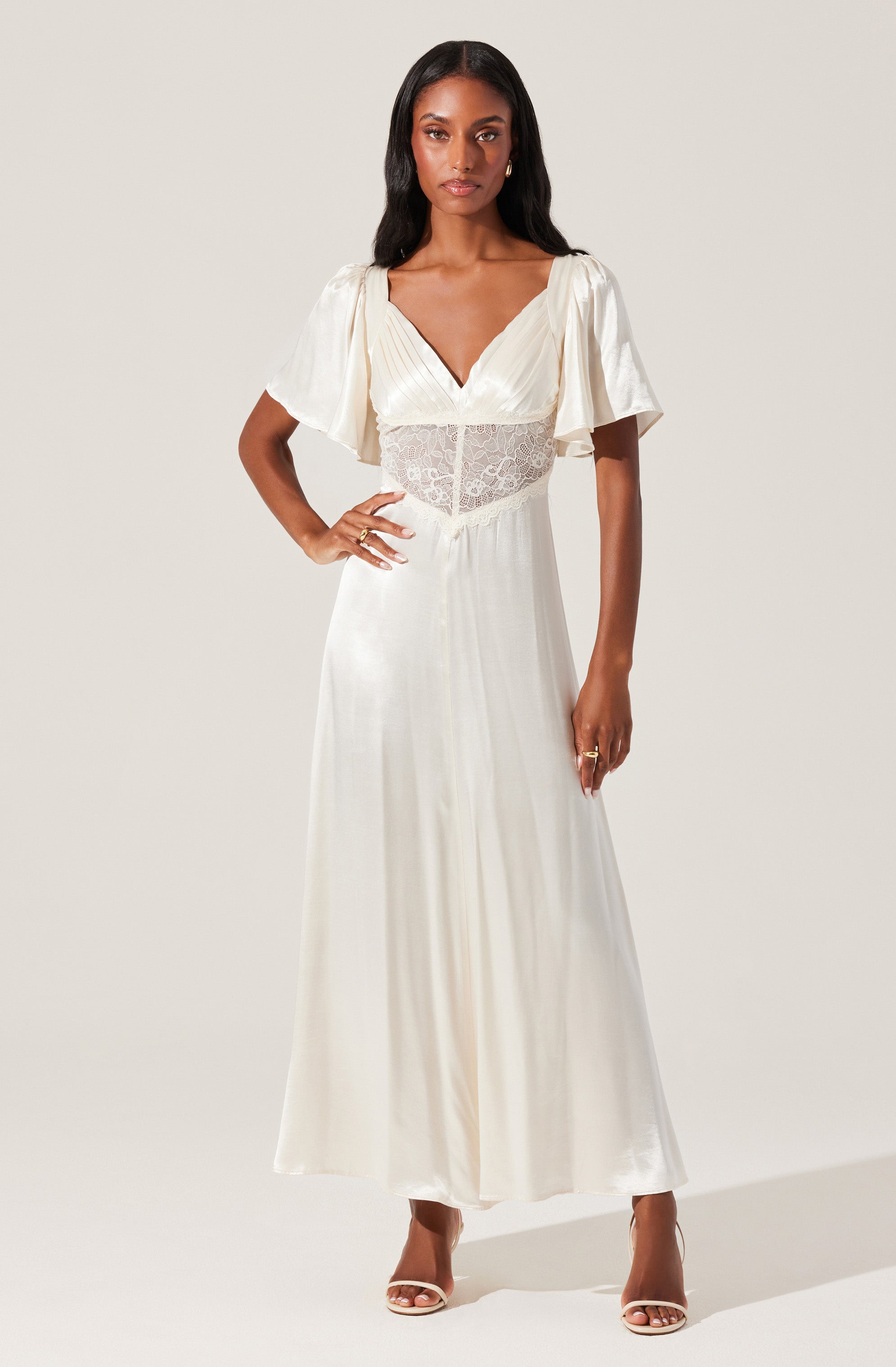 A woman is wearing a long, elegant white dress with short, puffed sleeves made of a flowing, silky fabric. The dress features a V-neckline and a sheer lace panel around the waist, adding a delicate touch. She is also wearing high-heeled shoes and accessorized with rings and earrings.