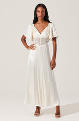 A woman is wearing a long, elegant white dress with short, puffed sleeves made of a flowing, silky fabric. The dress features a V-neckline and a sheer lace panel around the waist, adding a delicate touch. She is also wearing high-heeled shoes and accessorized with rings and earrings.