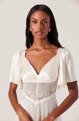 A woman is wearing an elegant cream-colored dress with a deep V neckline. The dress features puffed short sleeves made of shiny satin fabric and a fitted bodice with a lace panel at the waist, adding a touch of delicate detail. The dress has a gathered, flowing skirt.