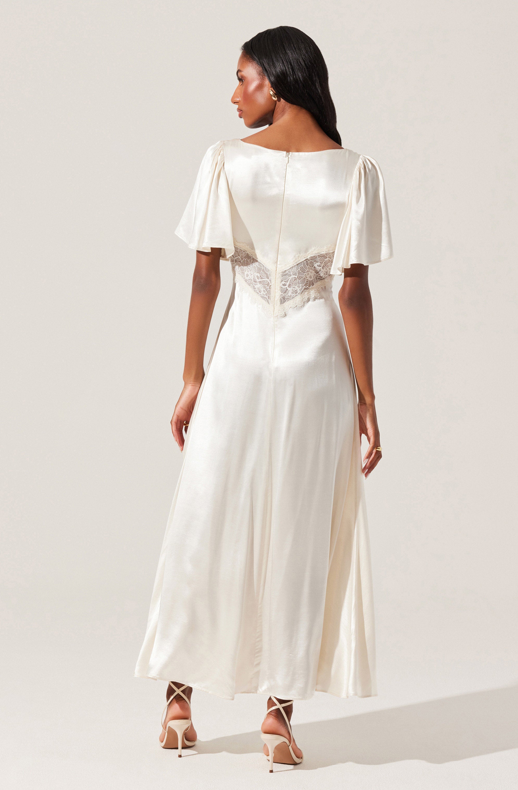 A woman is wearing an elegant, floor-length, ivory-colored satin dress with short puffed sleeves. The dress features a lace panel at the waist, adding a delicate touch. She completes her look with white high-heeled shoes that have ankle straps.