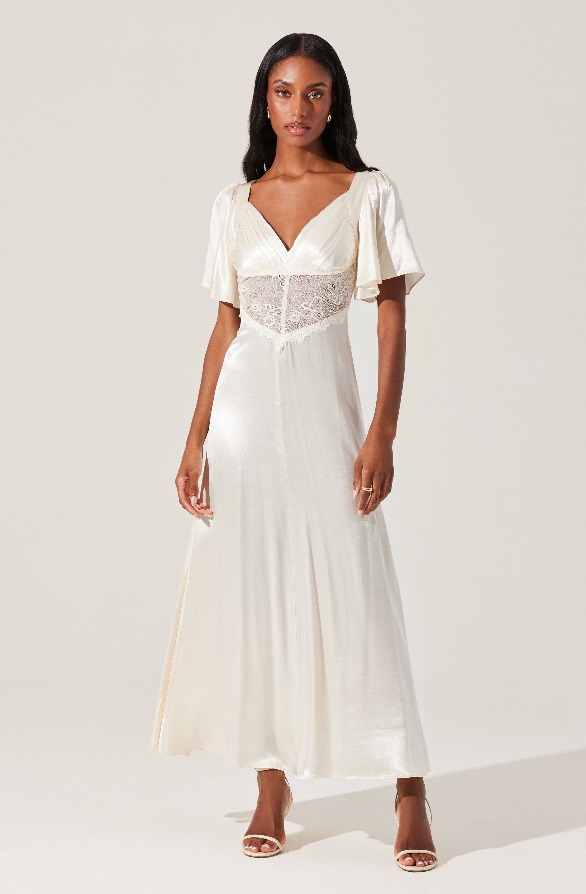 A woman is wearing a long, flowing white dress with a V-neckline and short, loose sleeves. The top part of the dress features a sheer lace panel around the waist, adding a delicate touch. She is also wearing open-toed high-heeled sandals.