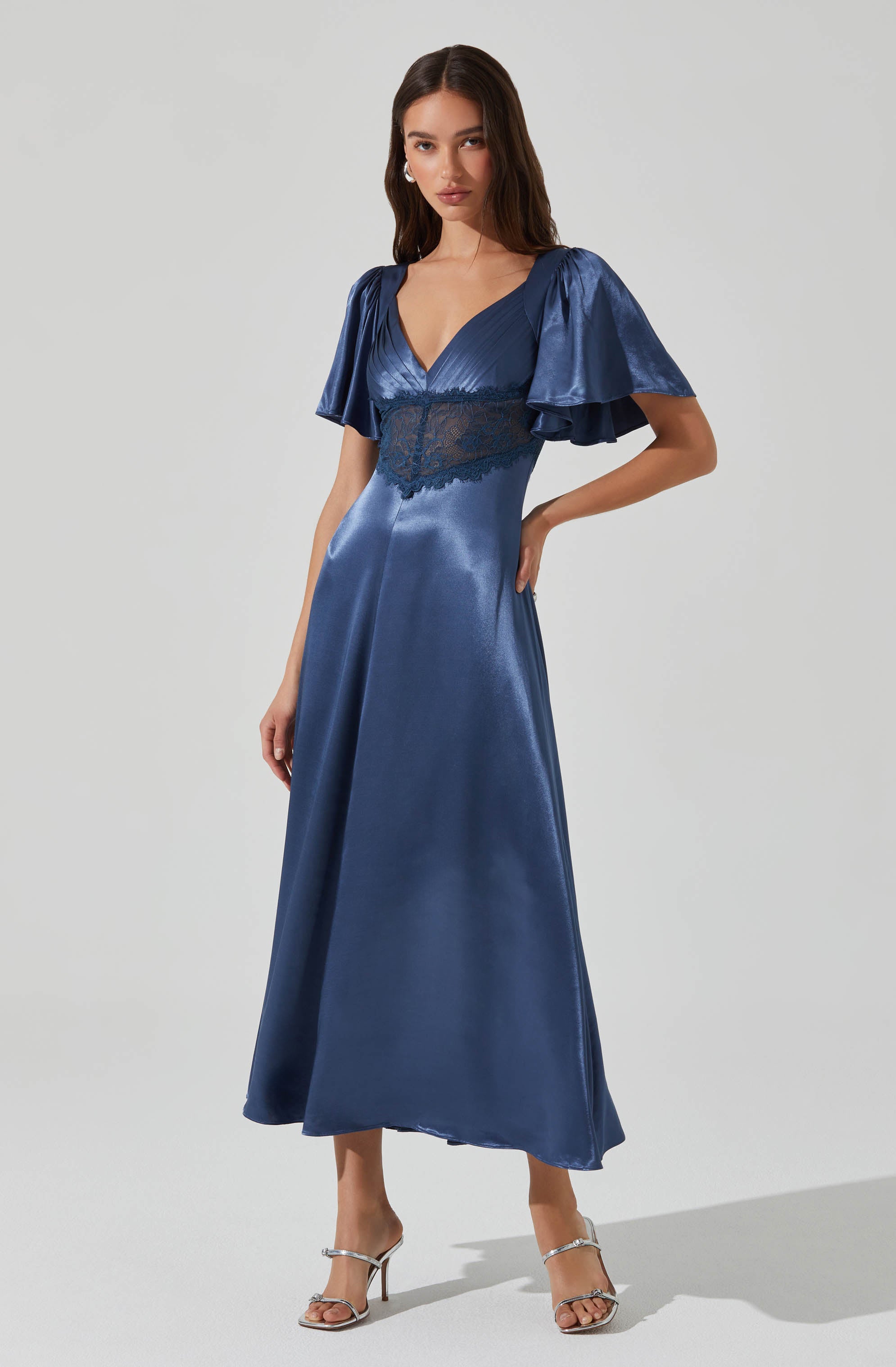 A woman is wearing a floor-length, shiny blue satin dress with a deep V neckline. The dress features short, puffy sleeves made of the same shiny blue satin fabric. There is a lace panel beneath the bust area that extends around the waist, adding a delicate detail to the dress. She is also wearing silver high-heeled sandals with thin straps.