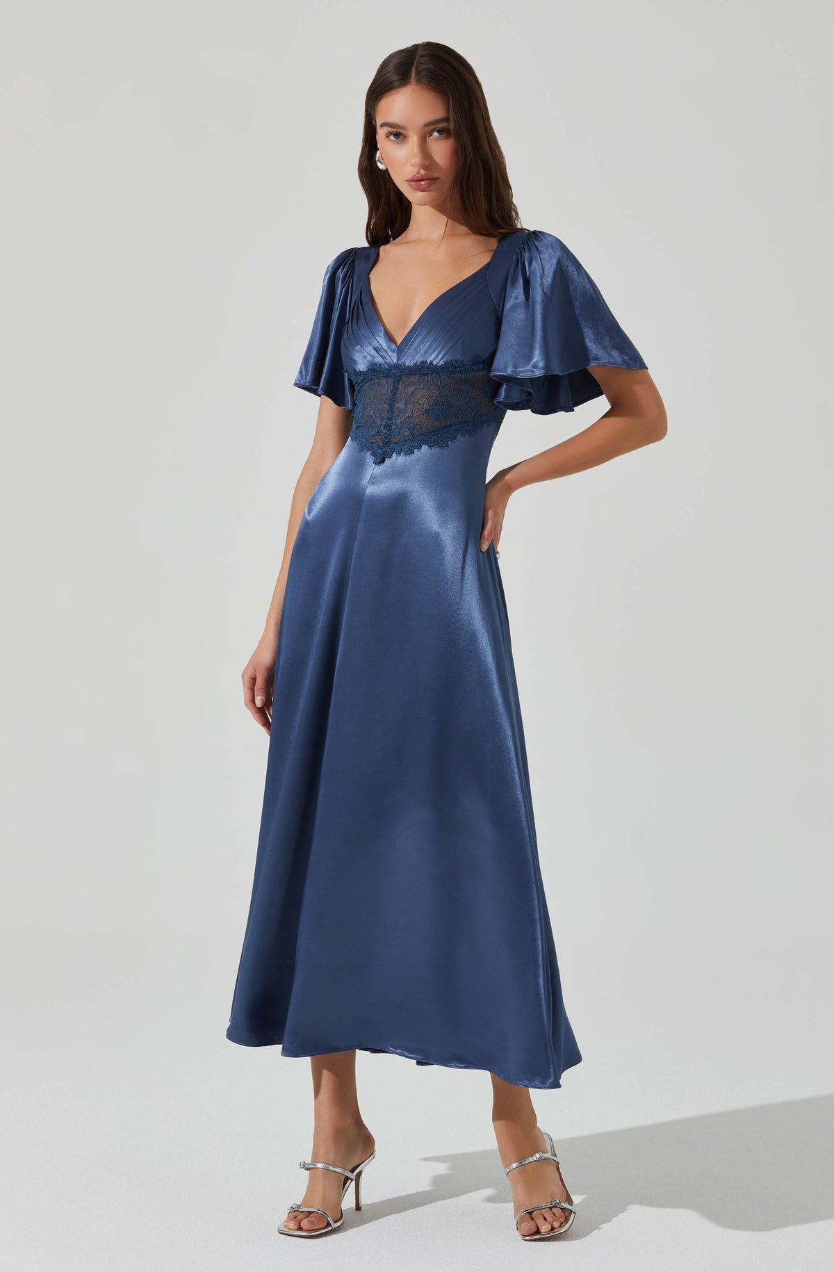 A woman is wearing a floor-length, shiny blue satin dress with a deep V neckline. The dress features short, puffy sleeves made of the same shiny blue satin fabric. There is a lace panel beneath the bust area that extends around the waist, adding a delicate detail to the dress. She is also wearing silver high-heeled sandals with thin straps.