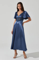A woman is wearing a floor-length, shiny blue satin dress with a deep V neckline. The dress features short, puffy sleeves made of the same shiny blue satin fabric. There is a lace panel beneath the bust area that extends around the waist, adding a delicate detail to the dress. She is also wearing silver high-heeled sandals with thin straps.