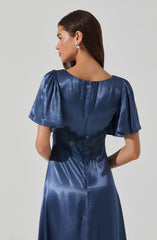 A woman is wearing a navy blue satin dress with short, puffed sleeves and a fitted waist. The dress features a lace panel at the back, adding a decorative touch. She has long, straight brown hair and is wearing silver hoop earrings.