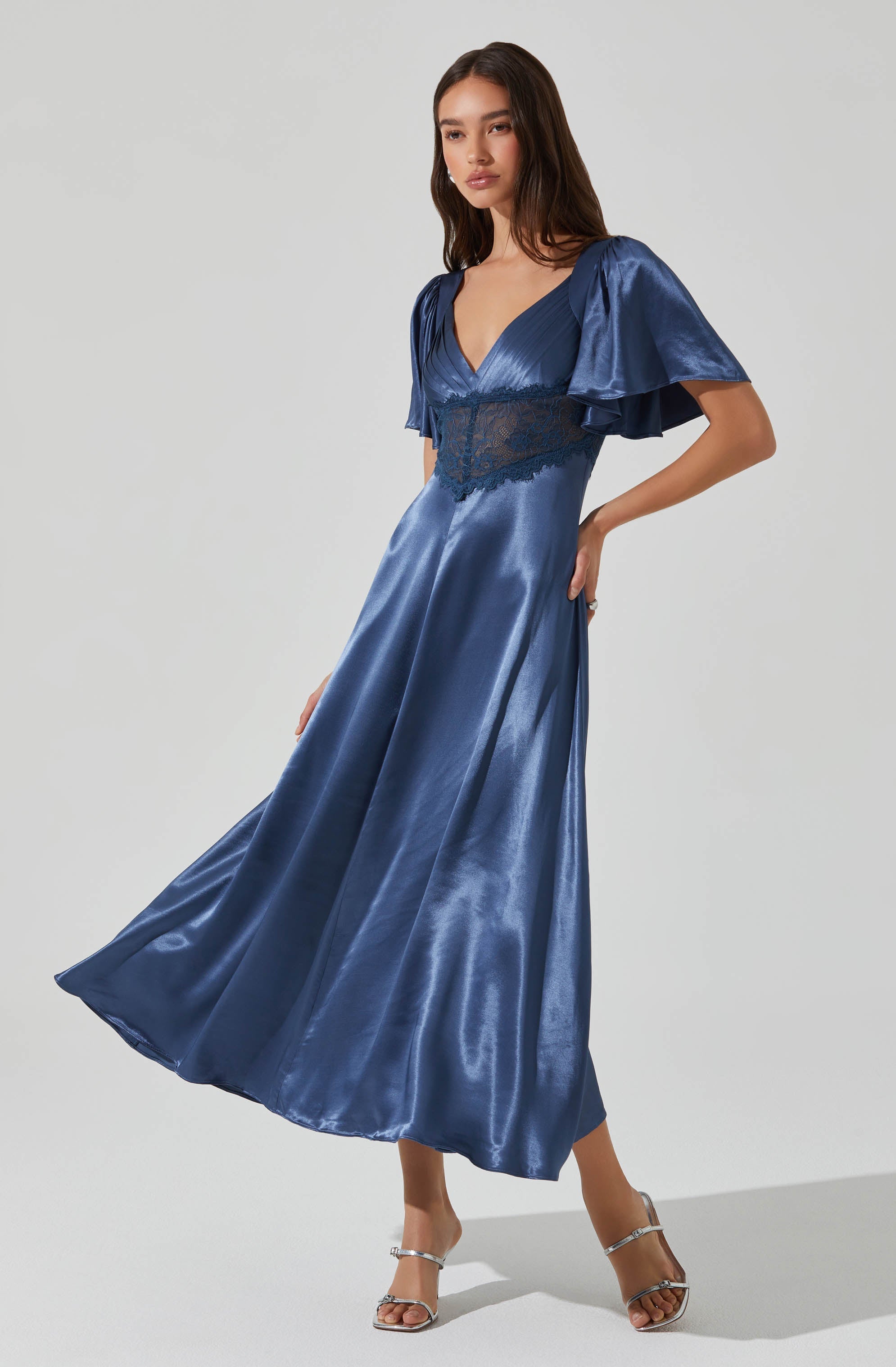 A woman is wearing a long, shiny, satin blue dress featuring a deep V neckline, puffed short sleeves with ruffles, and a waist detail with lace trim. She is also wearing high-heeled sandals.