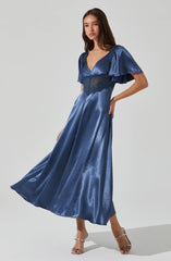 A woman is wearing a long, shiny, satin blue dress featuring a deep V neckline, puffed short sleeves with ruffles, and a waist detail with lace trim. She is also wearing high-heeled sandals.