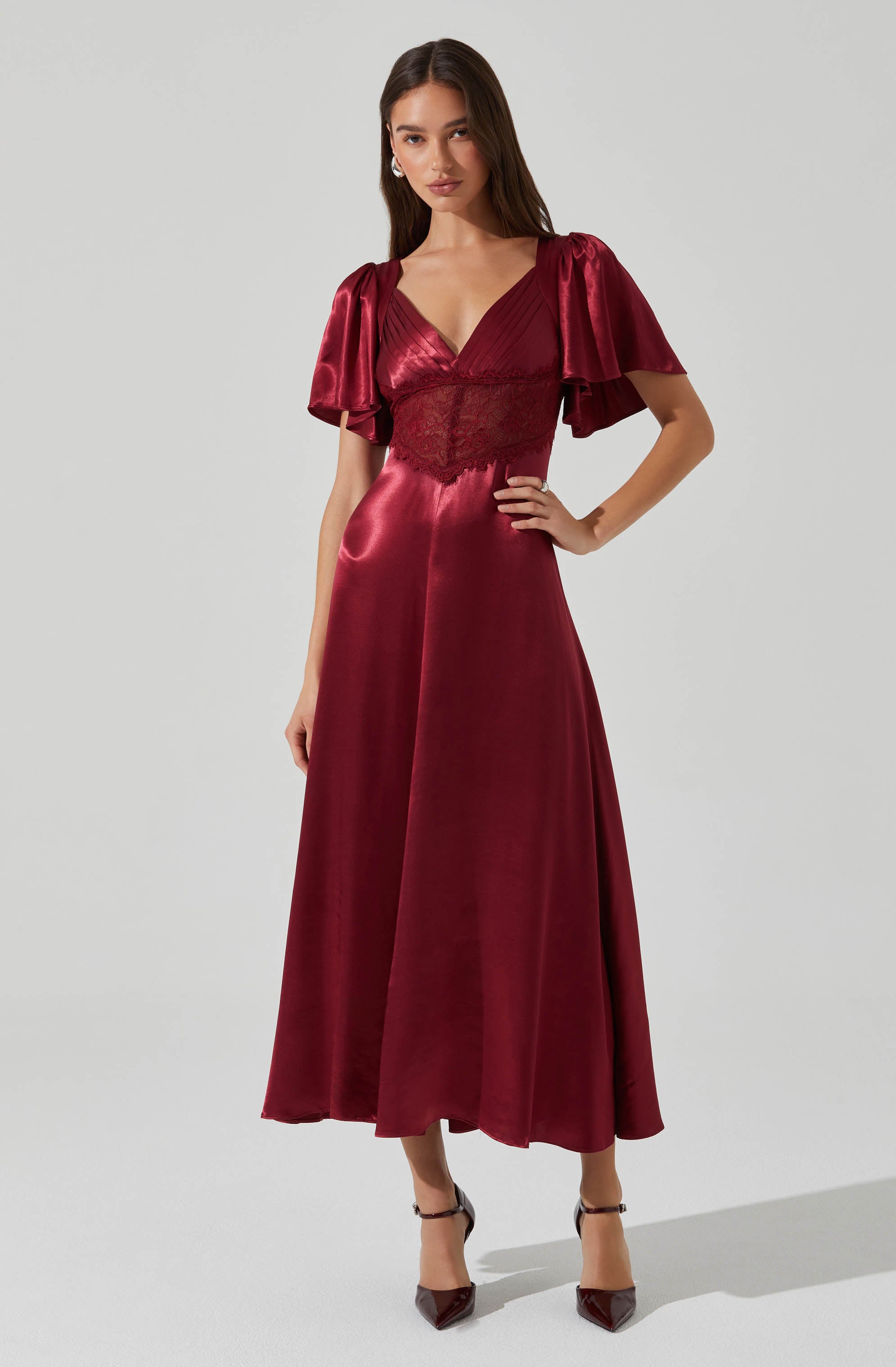 A woman is wearing a floor-length, deep red satin dress with a fitted bodice featuring lace details and a V-neckline. The dress has short, puffed sleeves made of a shiny satin fabric, contrasting with the lace waist. The skirt is long and flowing, made of the same satin material, creating an elegant appearance. She is also wearing pointed-toe high heels with ankle straps.