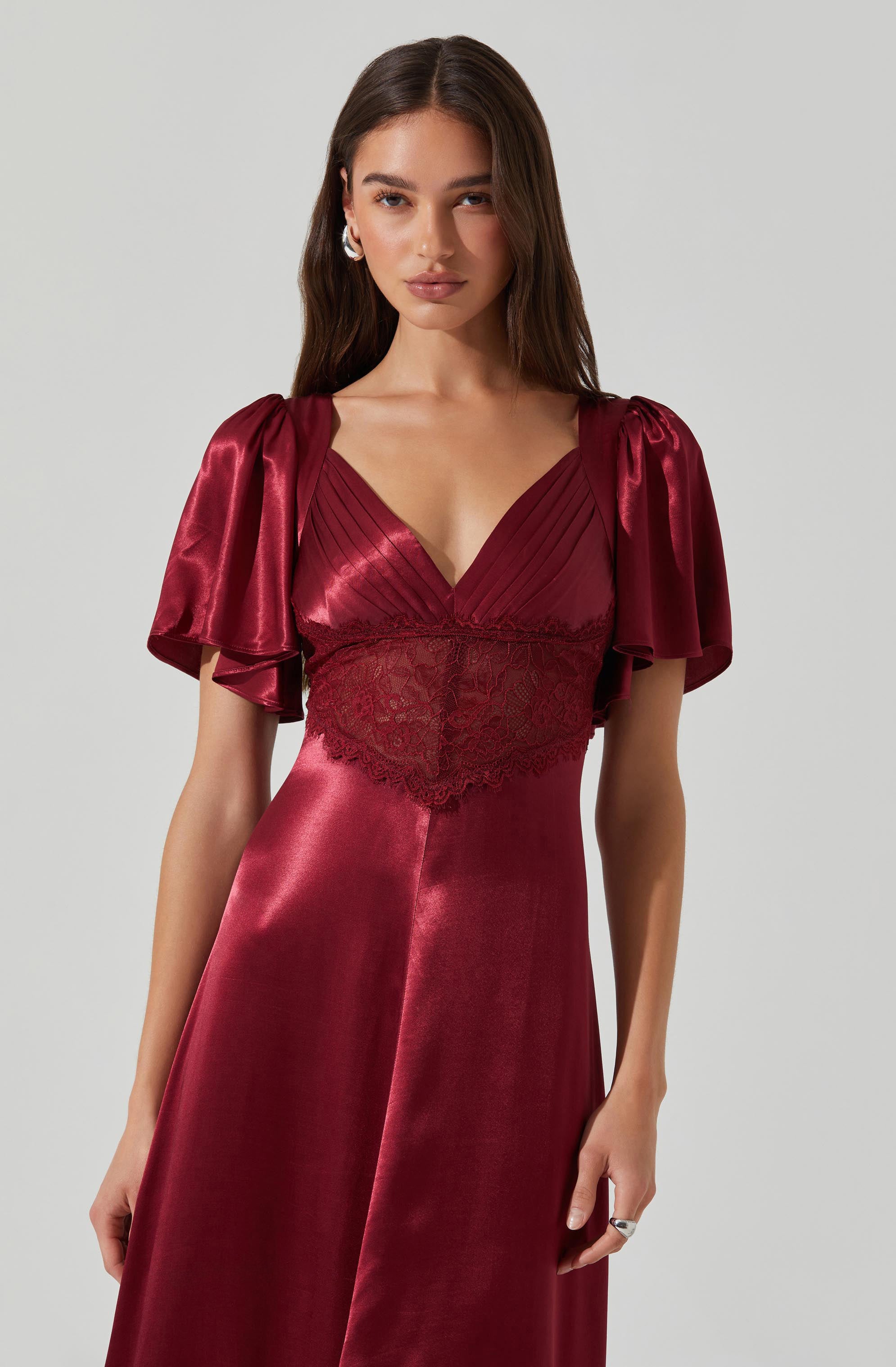 A woman is wearing a deep red, satin-like dress with a fitted bodice featuring pleated detailing and a lace panel at the waist. The dress has short, puffed sleeves made of shiny fabric that drape over her shoulders.